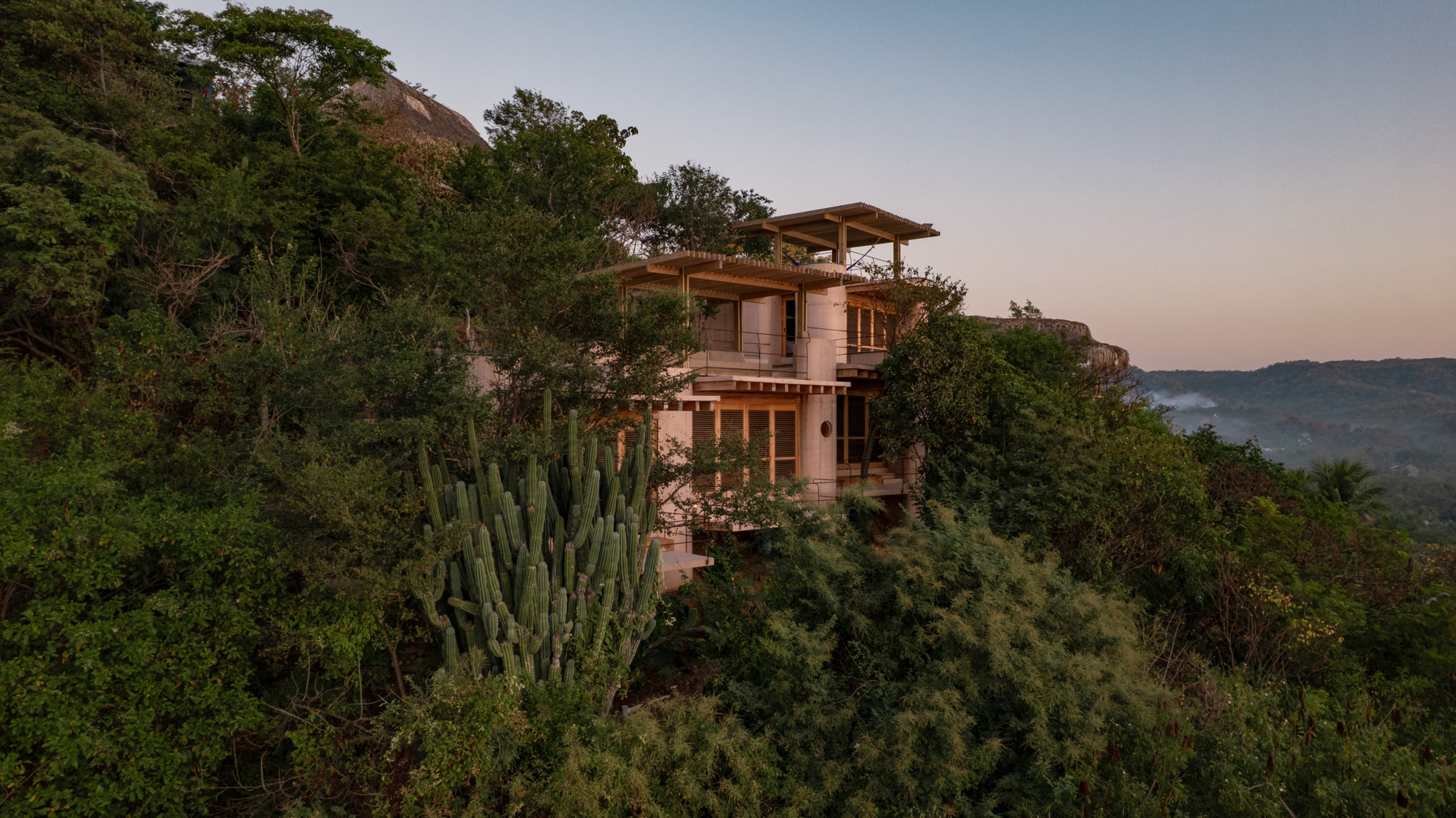Aerial view of the multi-level house with timber roof terraces, set among dense trees and a tall cactus at dusk