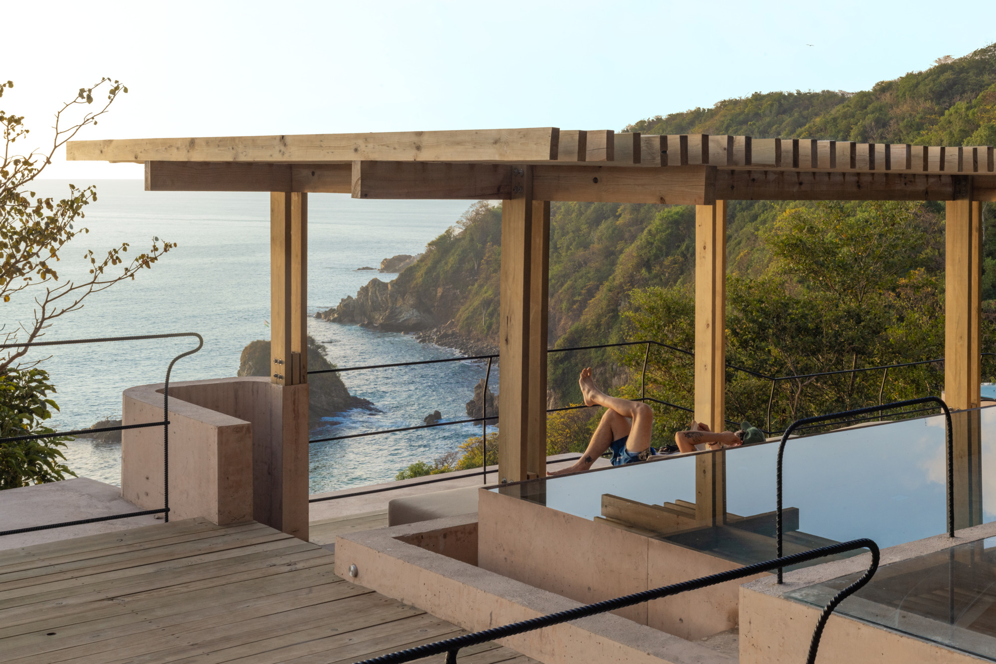 Timber pergola over a terrace with a plunge pool, where a person lounges facing a rocky coastline in low sun