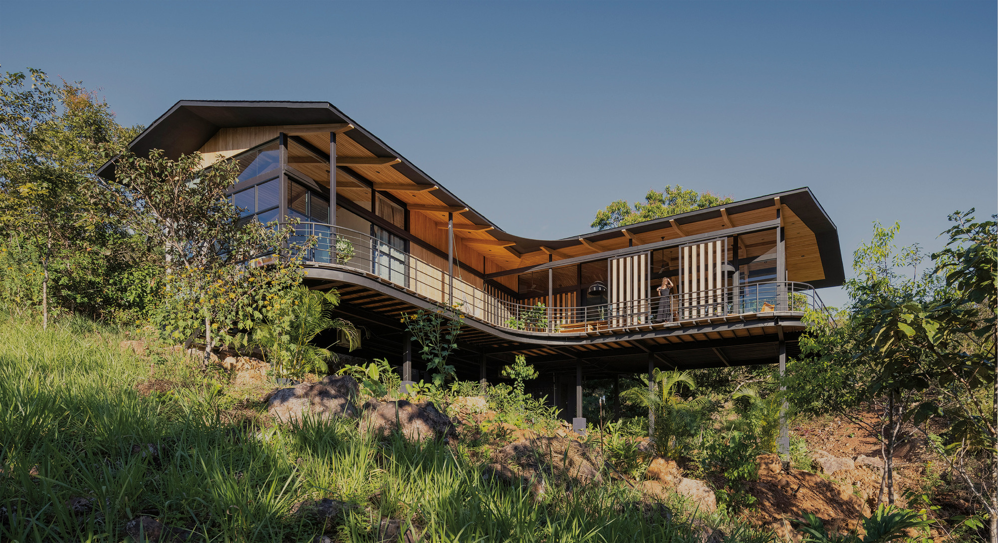 Wavy timber-soffit roof and curved balcony of a stilted house above a grassy, rocky slope under clear blue sky