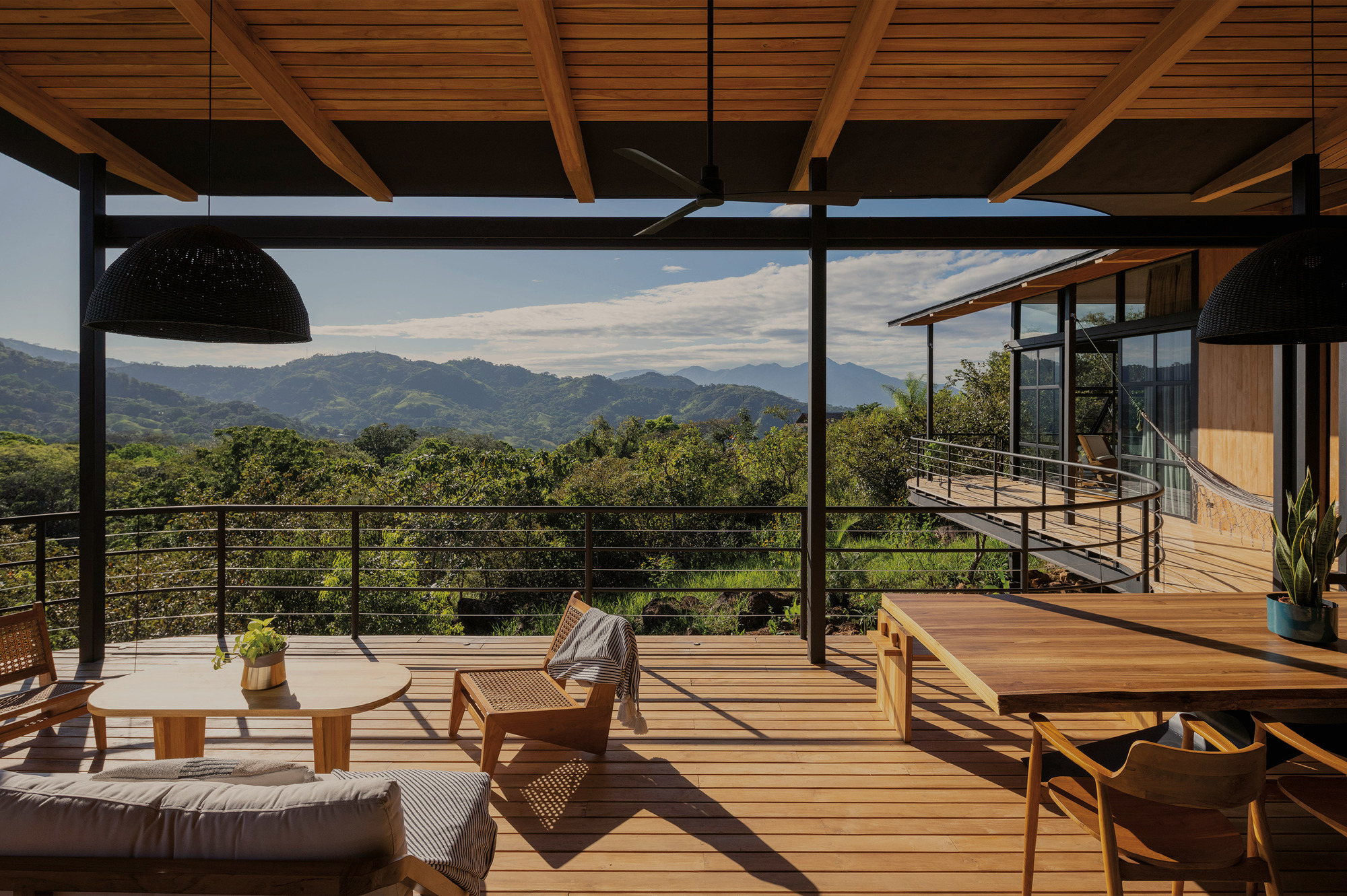 Covered timber deck with exposed beams, black pendant lamps and dining table facing forested mountains under scattered clouds