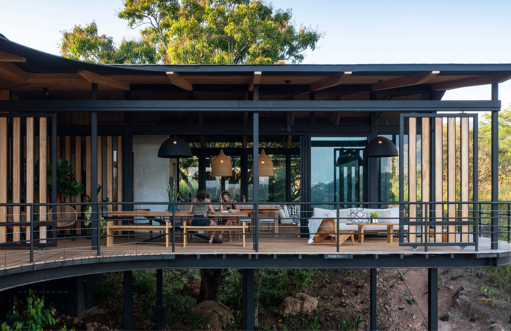 Curved elevated deck with steel railings and vertical timber slats, two people seated at a long dining table in daylight