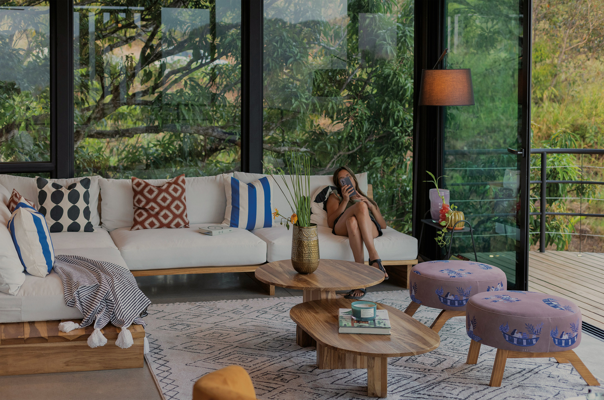 Living area behind floor-to-ceiling glass with a white sofa, wooden coffee tables, and a woman looking at her phone among tree reflections