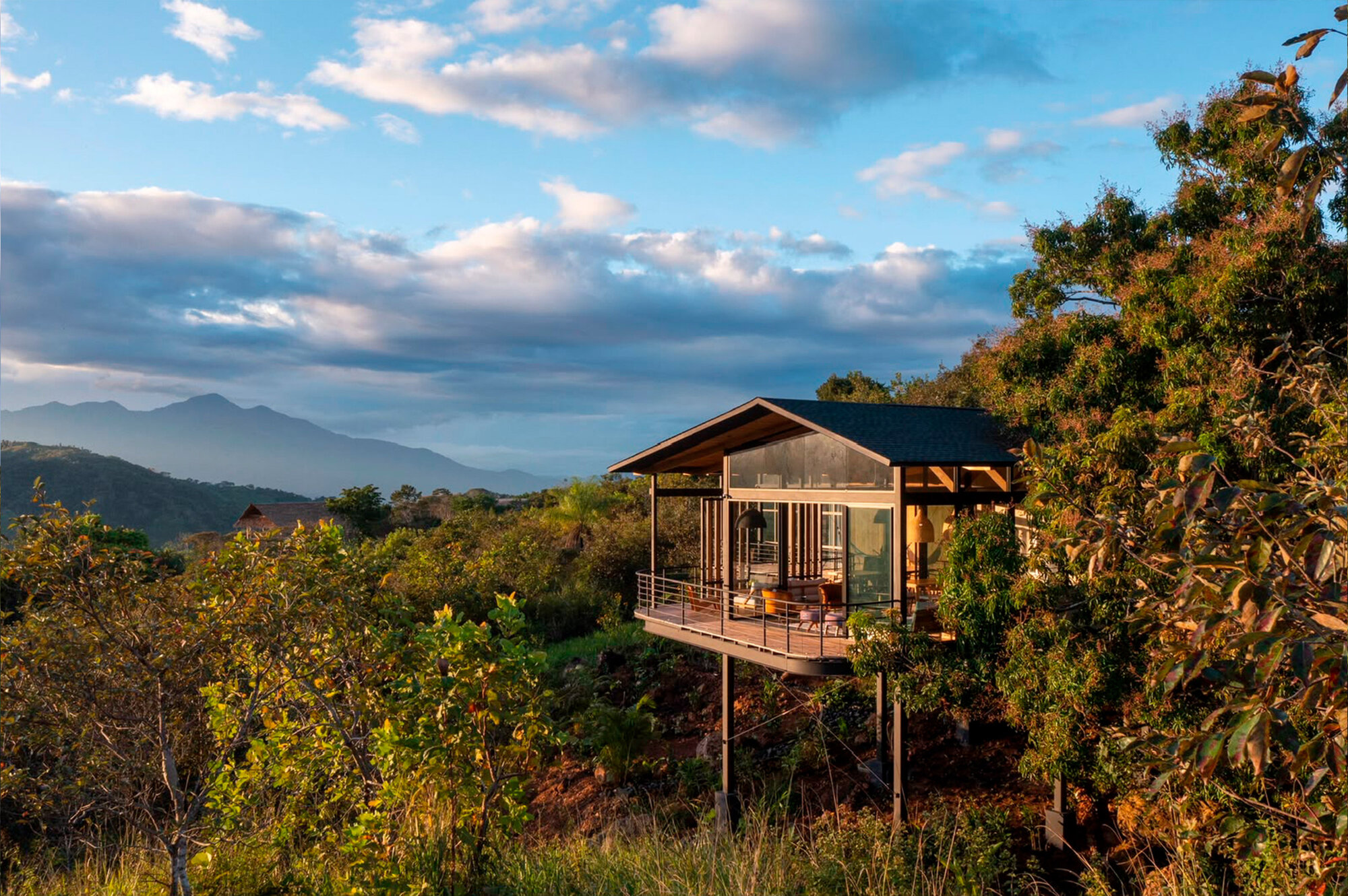 Glazed timber pavilion on slender steel stilts above a forested slope, lit by low sun with mountains and clouds behind