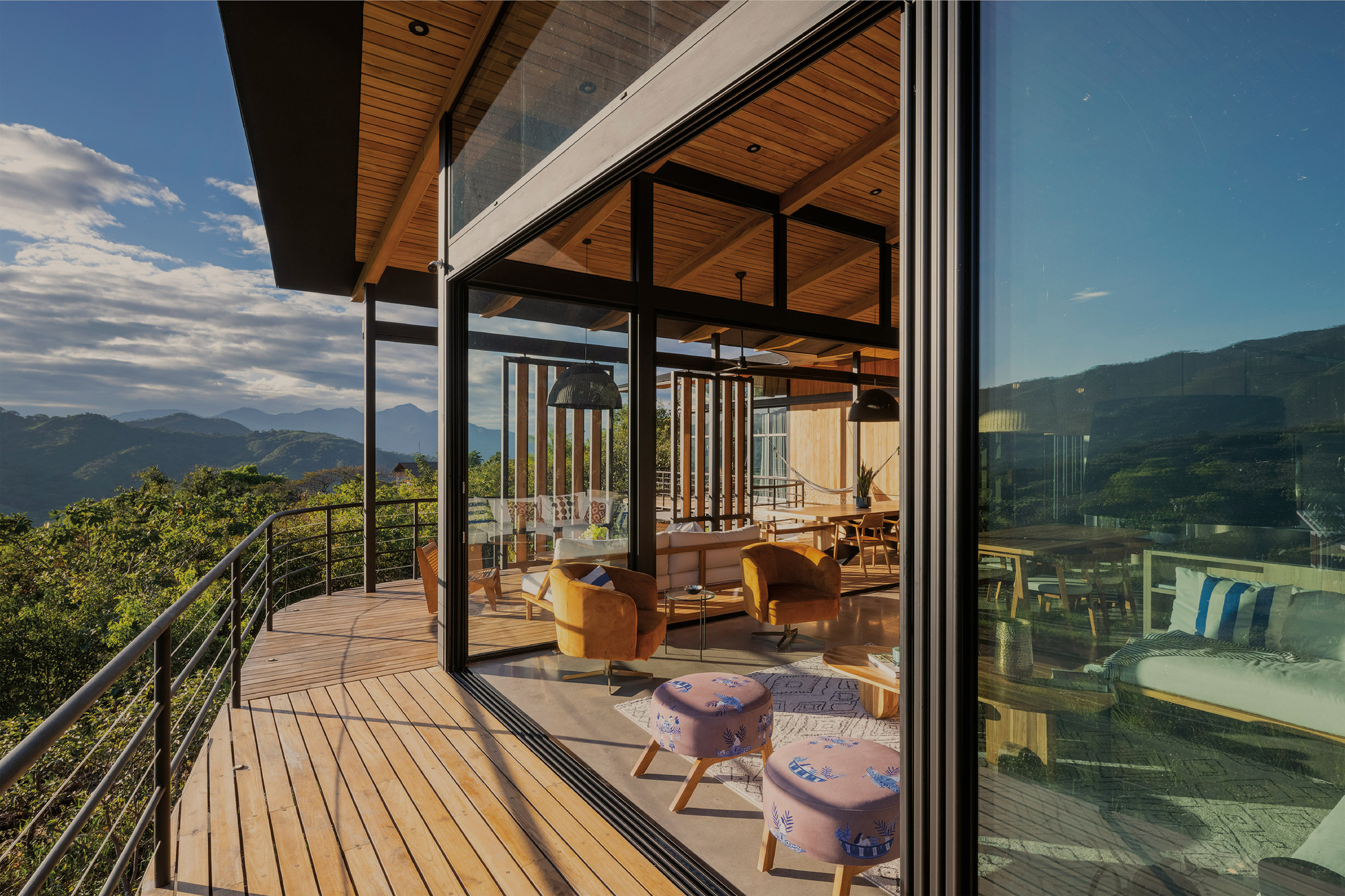 Corner of the sliding glass wall opening a living room to a wood deck with steel railing and mountain view
