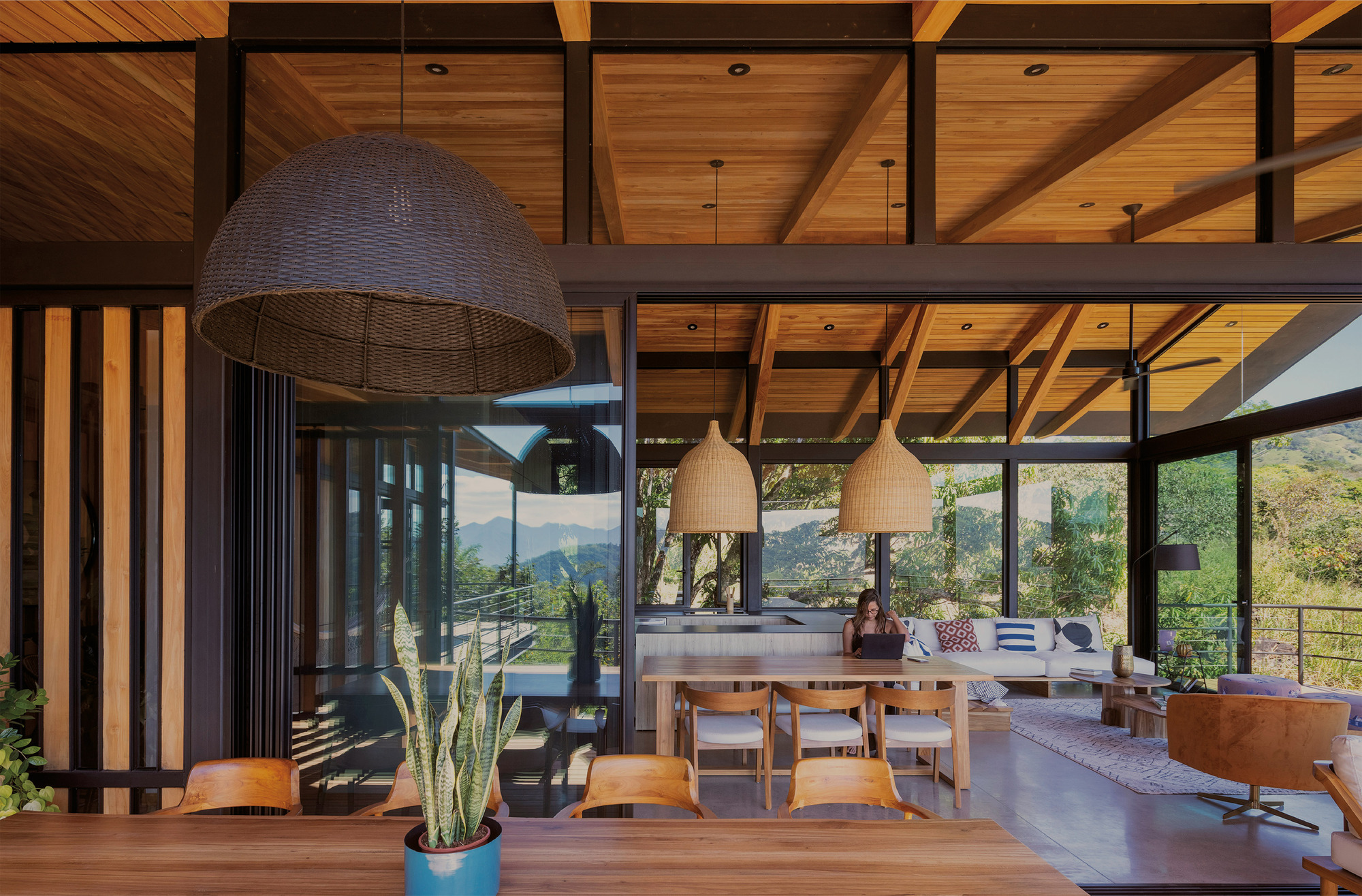 Dining table and woven pendant lamps beneath exposed timber rafters, with a person working on a laptop in the lounge beyond