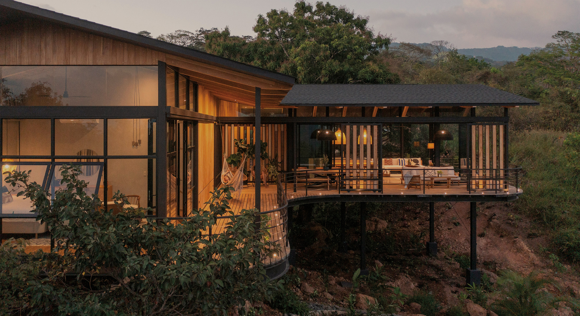 Dusk view of the elevated wood and glass house on steel posts, its lit rooms glowing above a rocky hillside