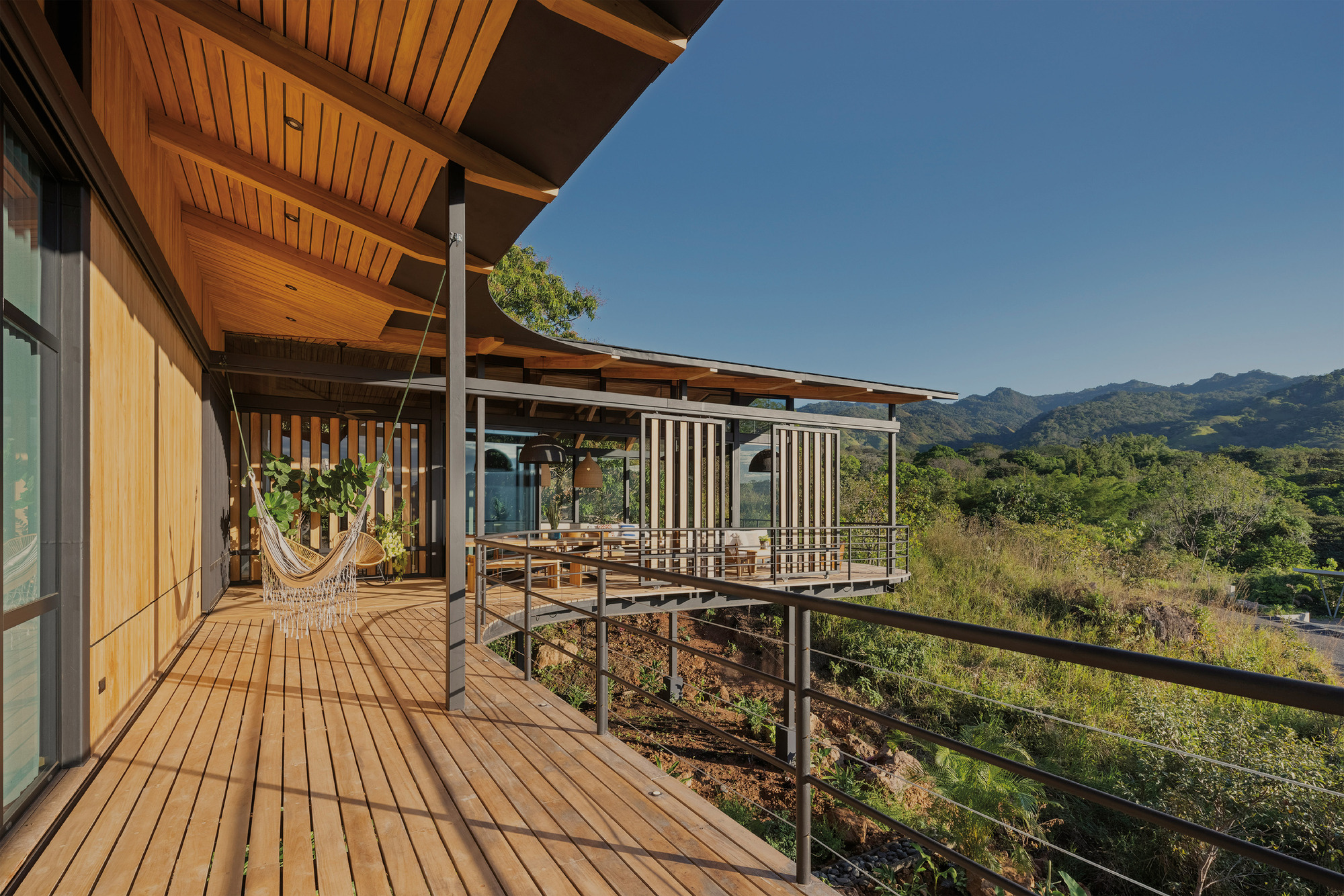 Long timber deck with a hammock and steel cable railing curving along the house under a clear blue sky