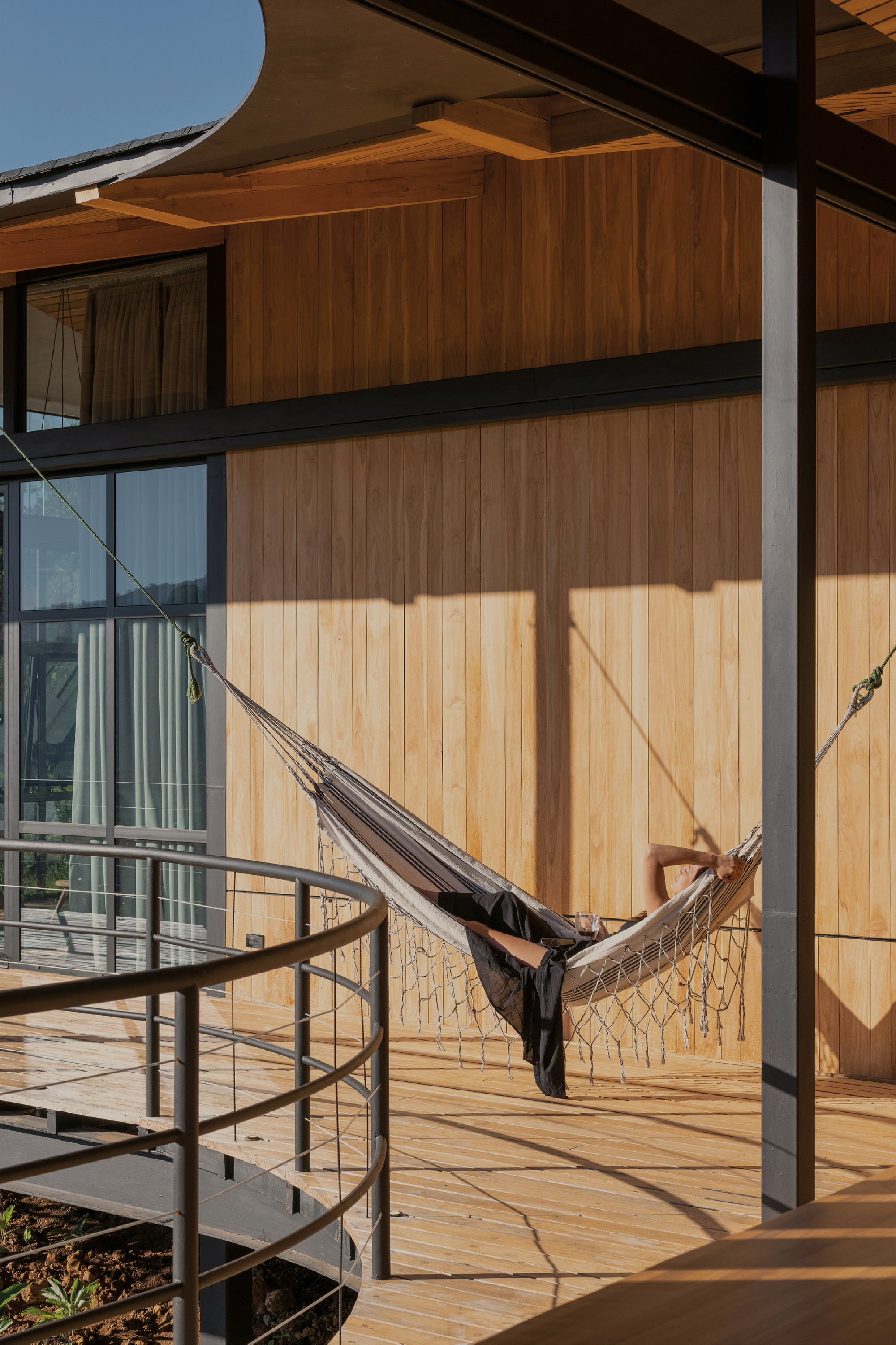 Person resting in a fringed hammock on a timber deck beside a curved steel railing in low sunlight