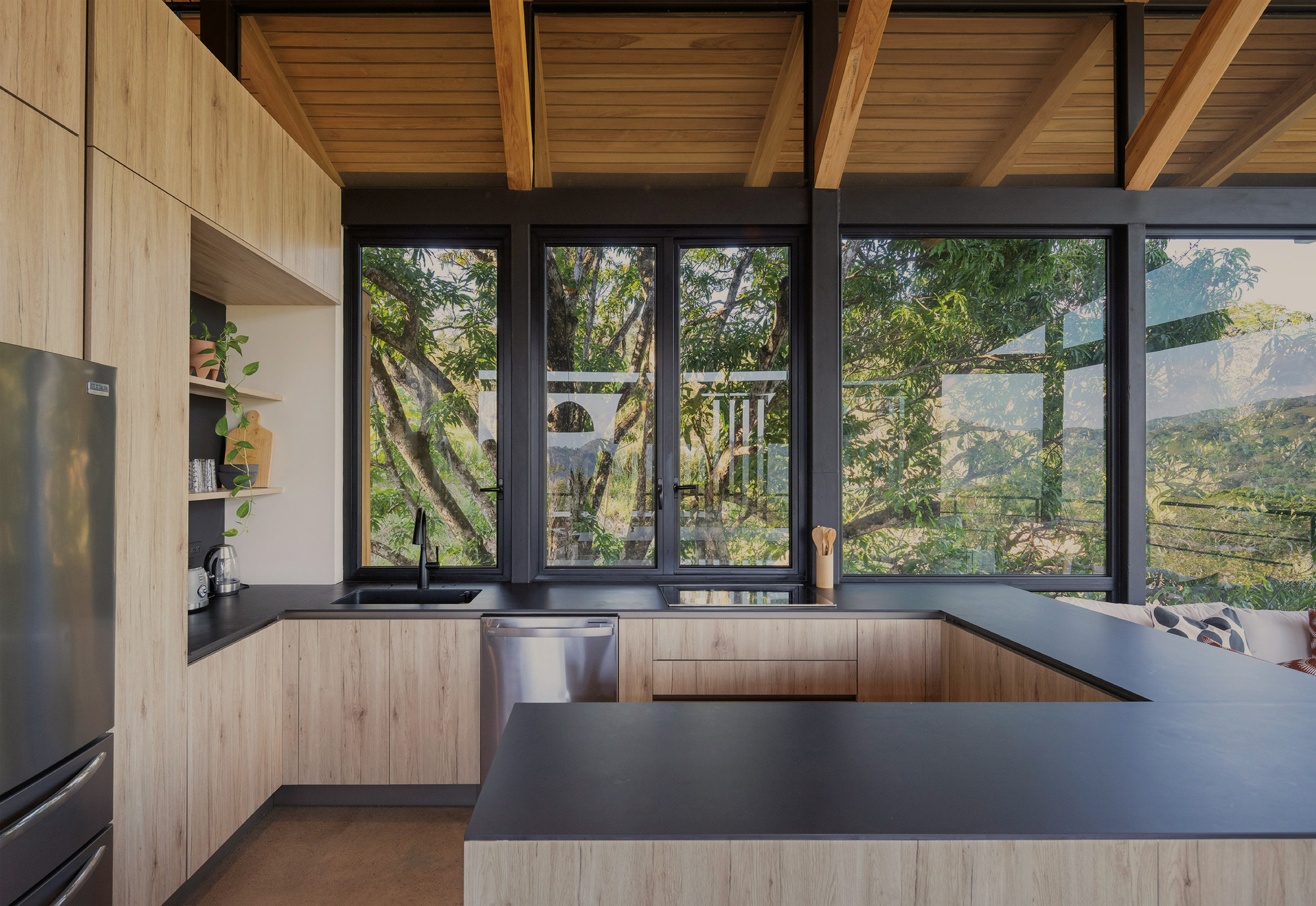 Kitchen with pale wood cabinets and a dark counter, framed by black windows looking into leafy tree canopy