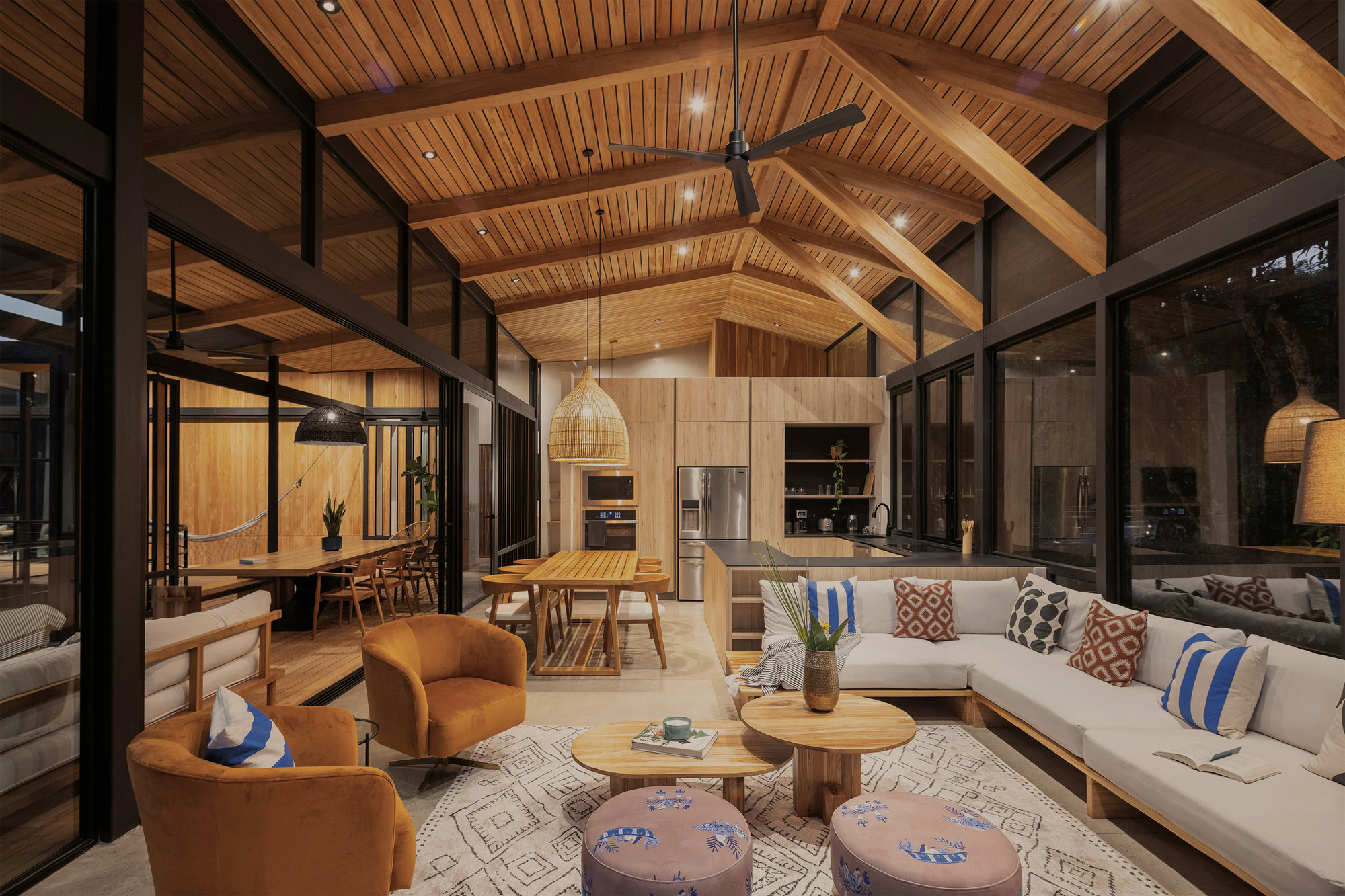 Open living area with exposed timber roof beams, white sofa and woven pendant lamps at dusk