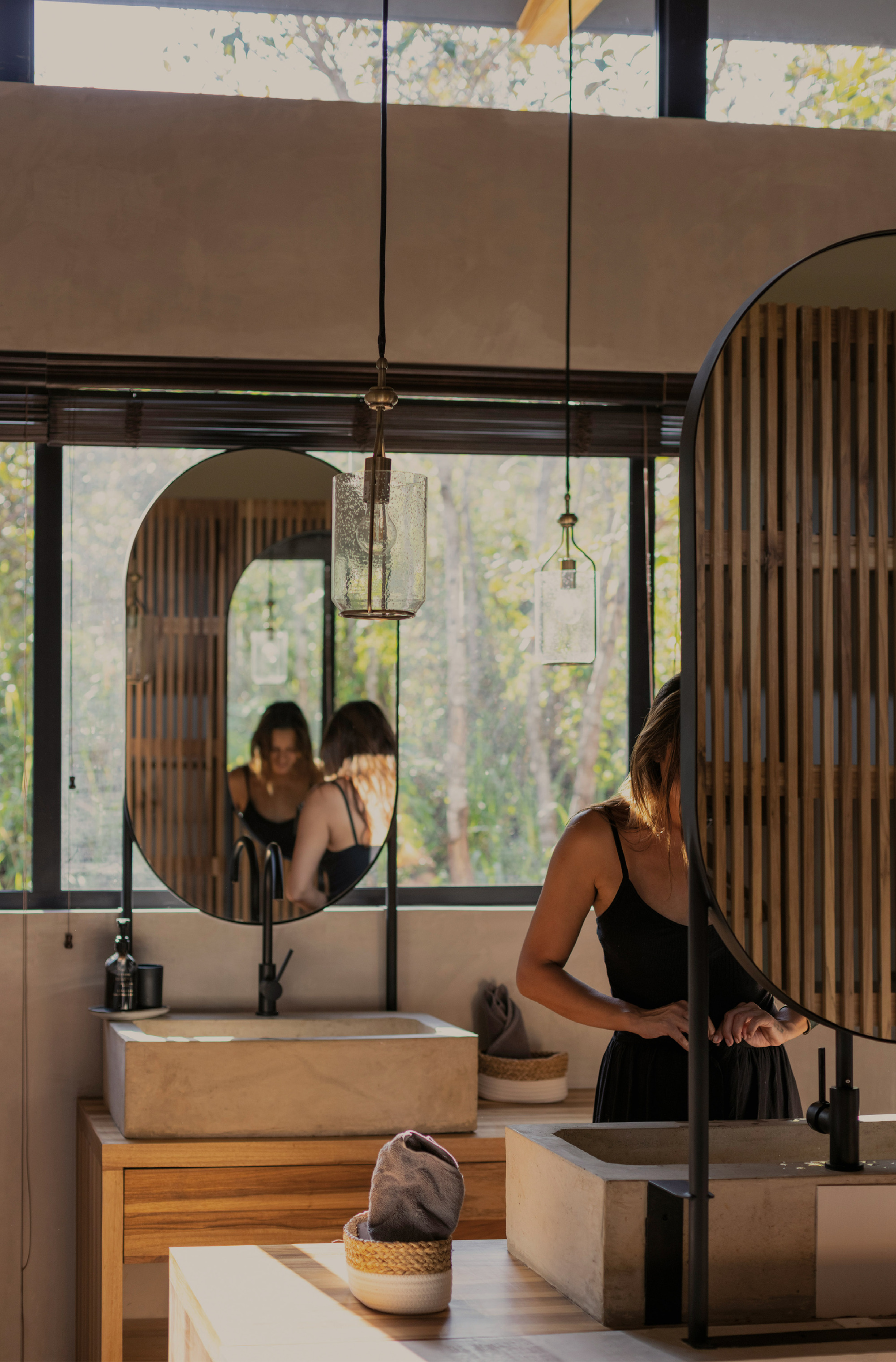 Bathroom with two stone basins and oval mirrors reflecting a woman at the sink against forest light