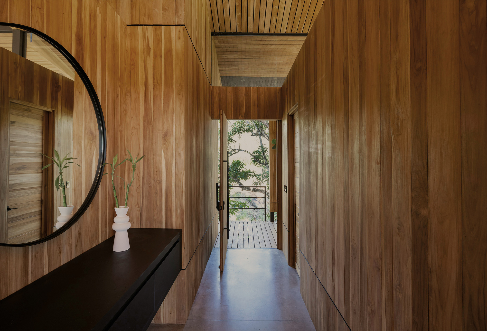 Timber-clad corridor with a round mirror and black floating console leading to an open door and deck