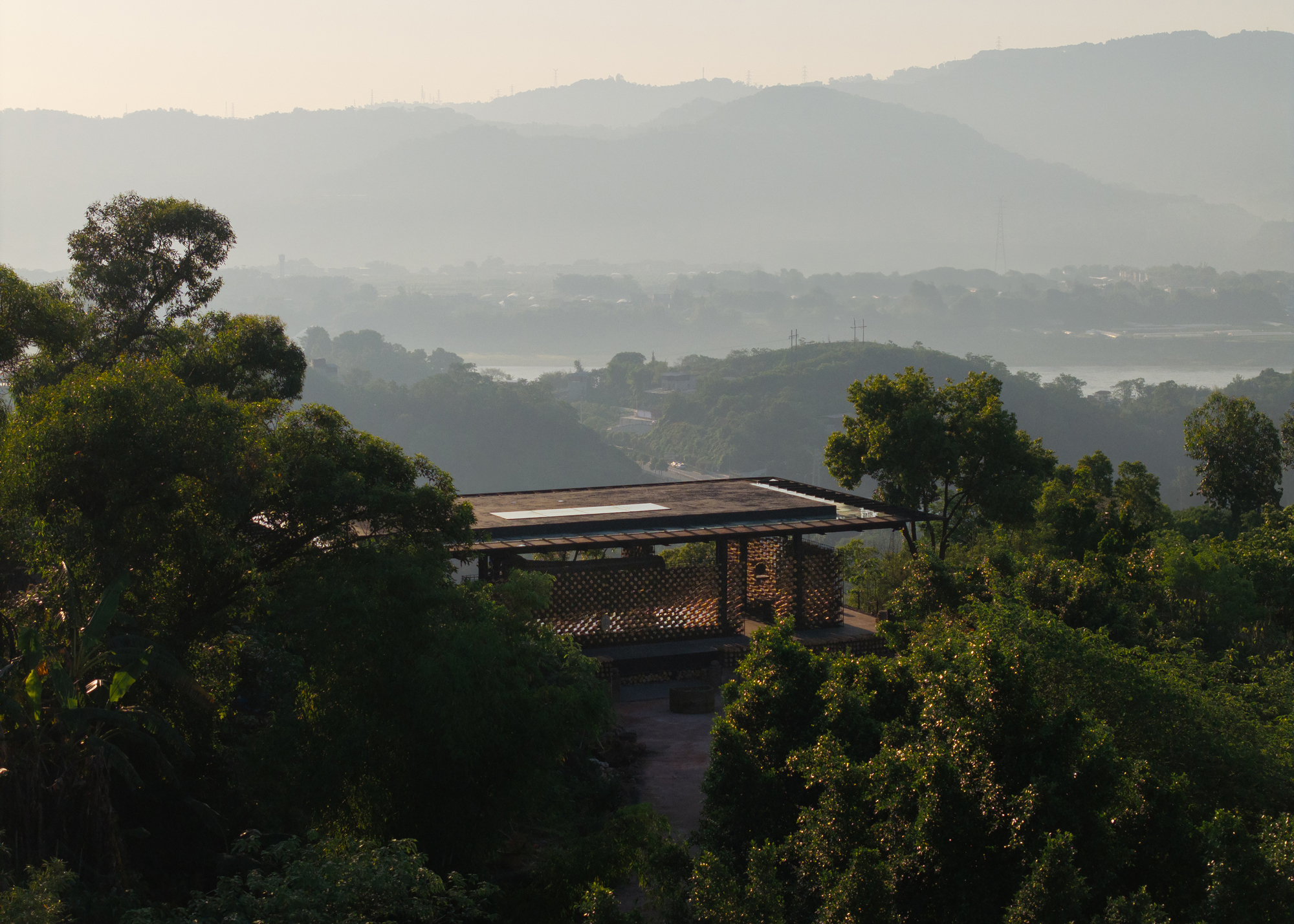 Flat-roofed pavilion with a lattice screen wall perched among treetops above hazy layered hills in morning light