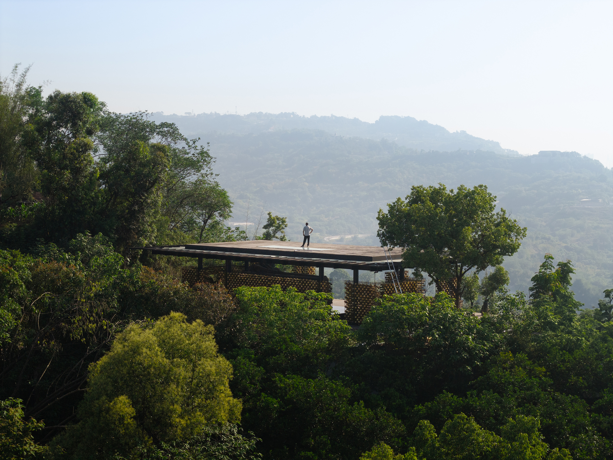 Person standing on the thin flat roof of the pavilion, framed by dense green canopy and distant ridges