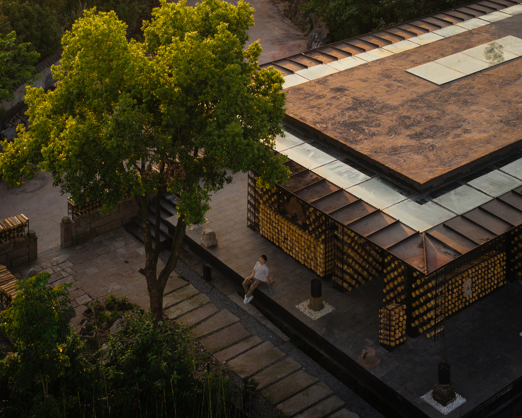 Aerial view of the pavilion's skylit roof and log-filled walls, with a seated visitor beside a leafy tree at golden hour