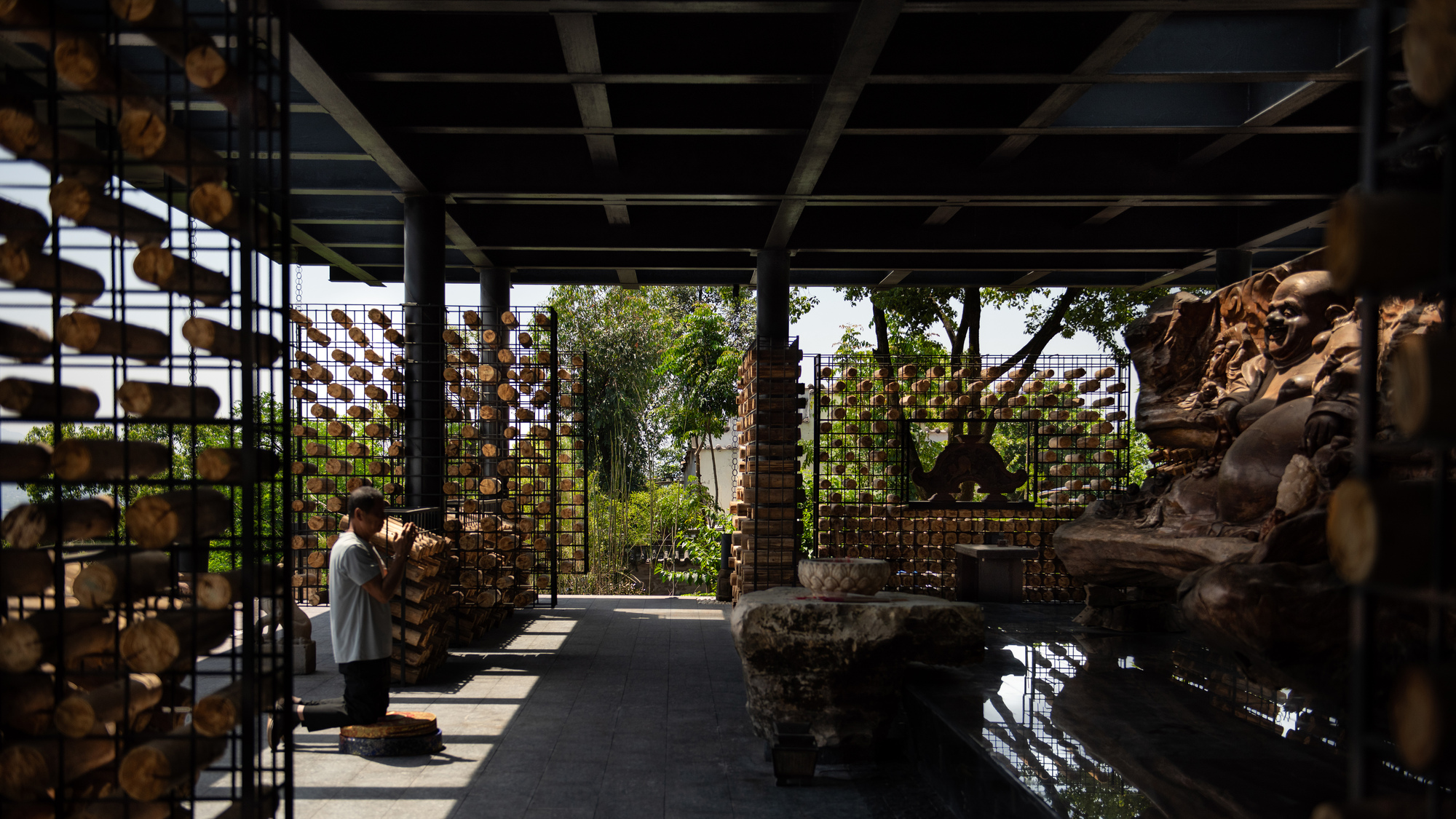 Open interior hall with steel columns and log-filled cages, a carved stone figure on the right and a worker stacking wood in dappled light
