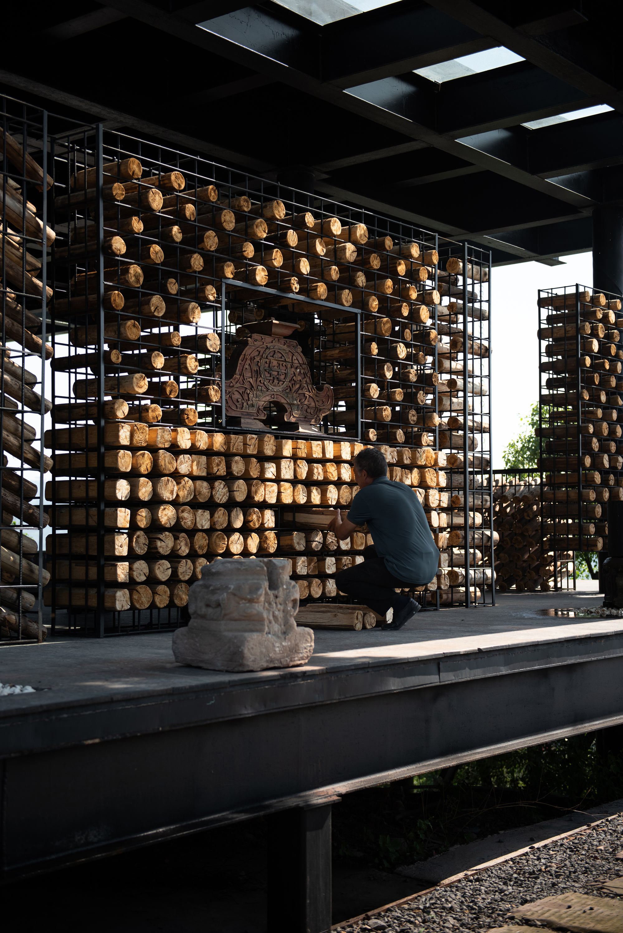 Worker kneeling to stack cut logs inside a steel grid frame around a carved stone ornament, with a stone fragment in the foreground