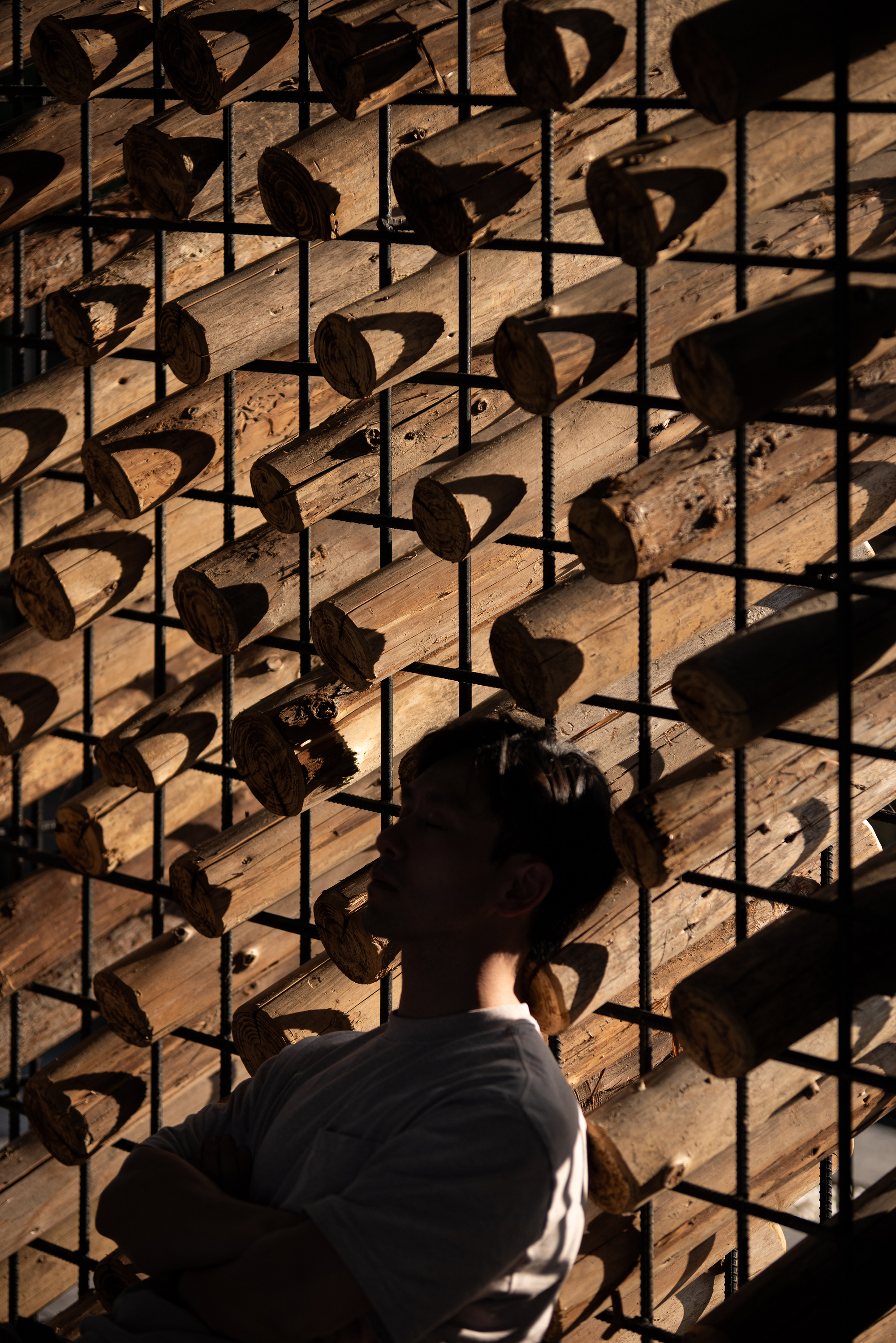 Close-up of a man in profile behind a steel rebar grid holding angled log ends, sunlight casting sharp shadows