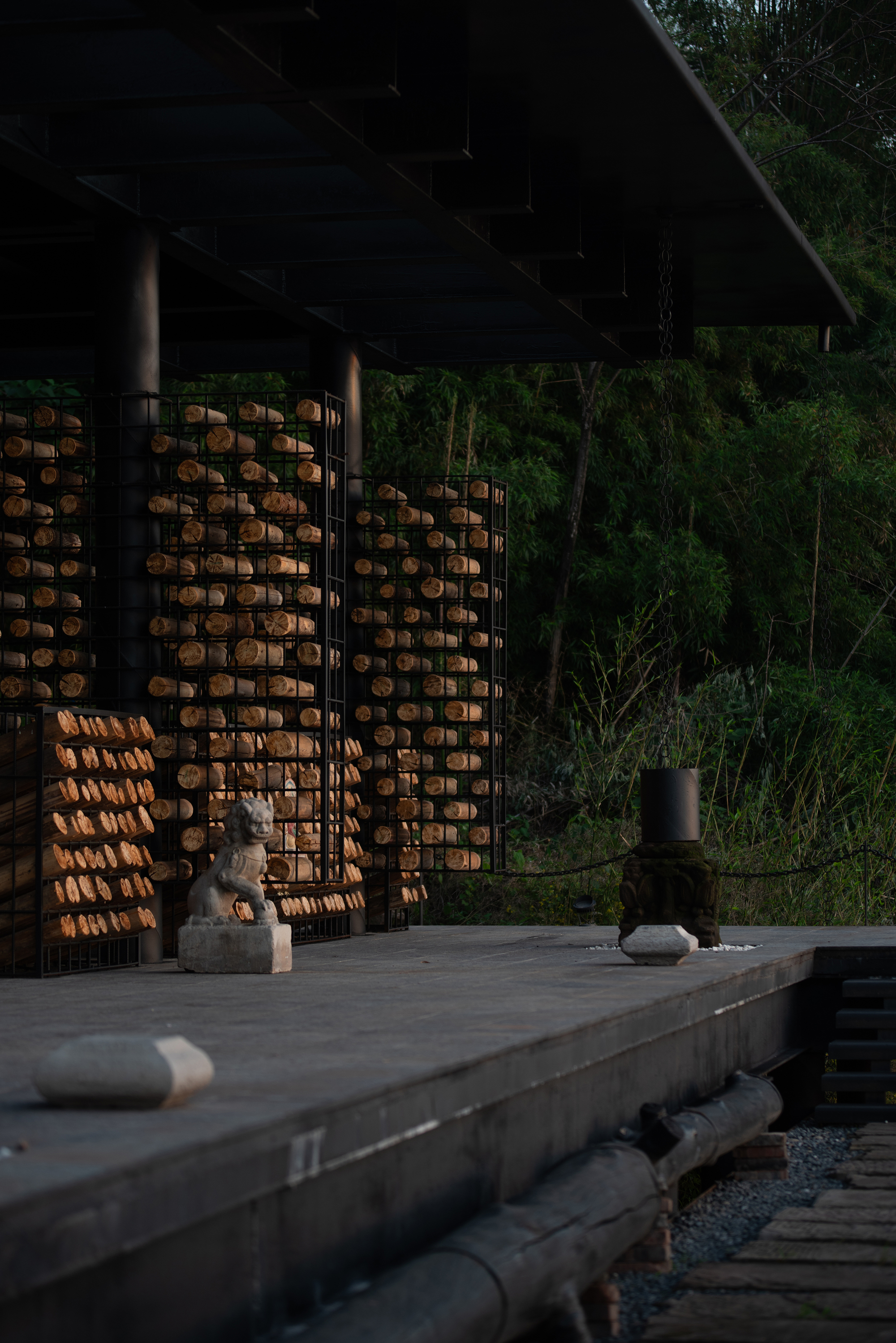 Stone lion statue on a dark concrete platform beside cages of stacked log ends, under a steel roof at dusk