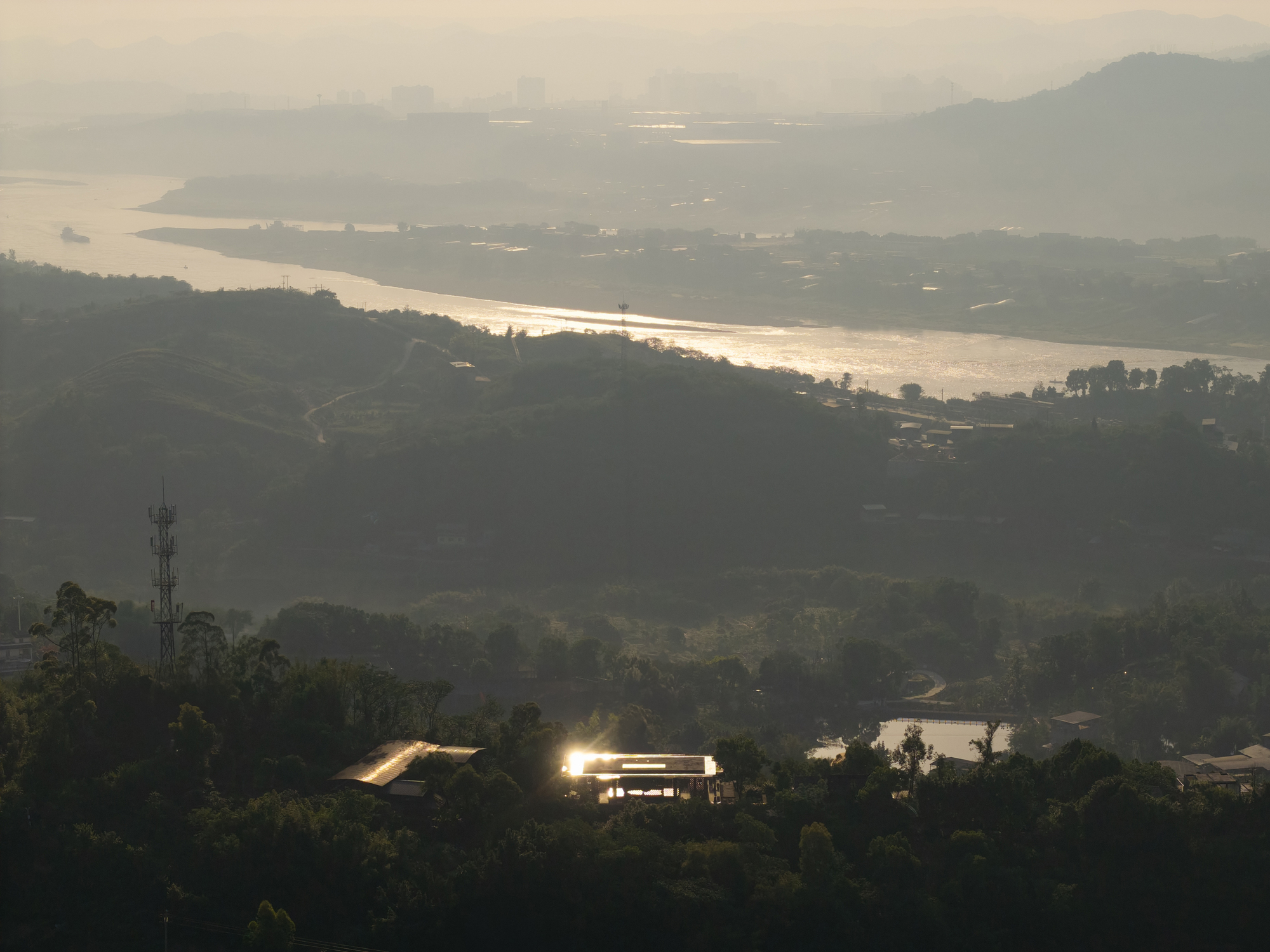 Distant view of the pavilion roof glinting among forested hills above a river in afternoon haze