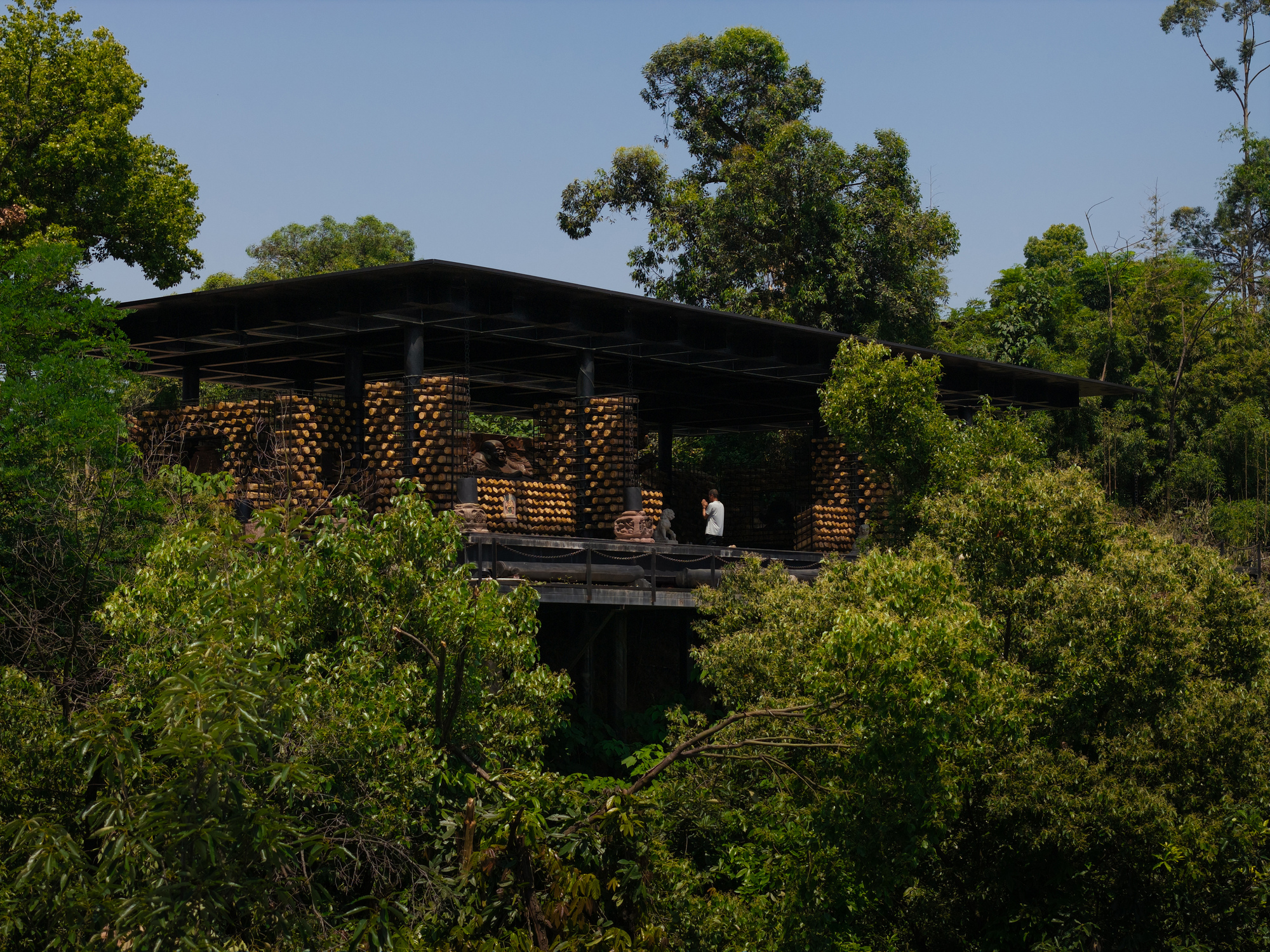 Pavilion with a flat dark roof and log-filled walls seen through dense green trees under a clear blue sky