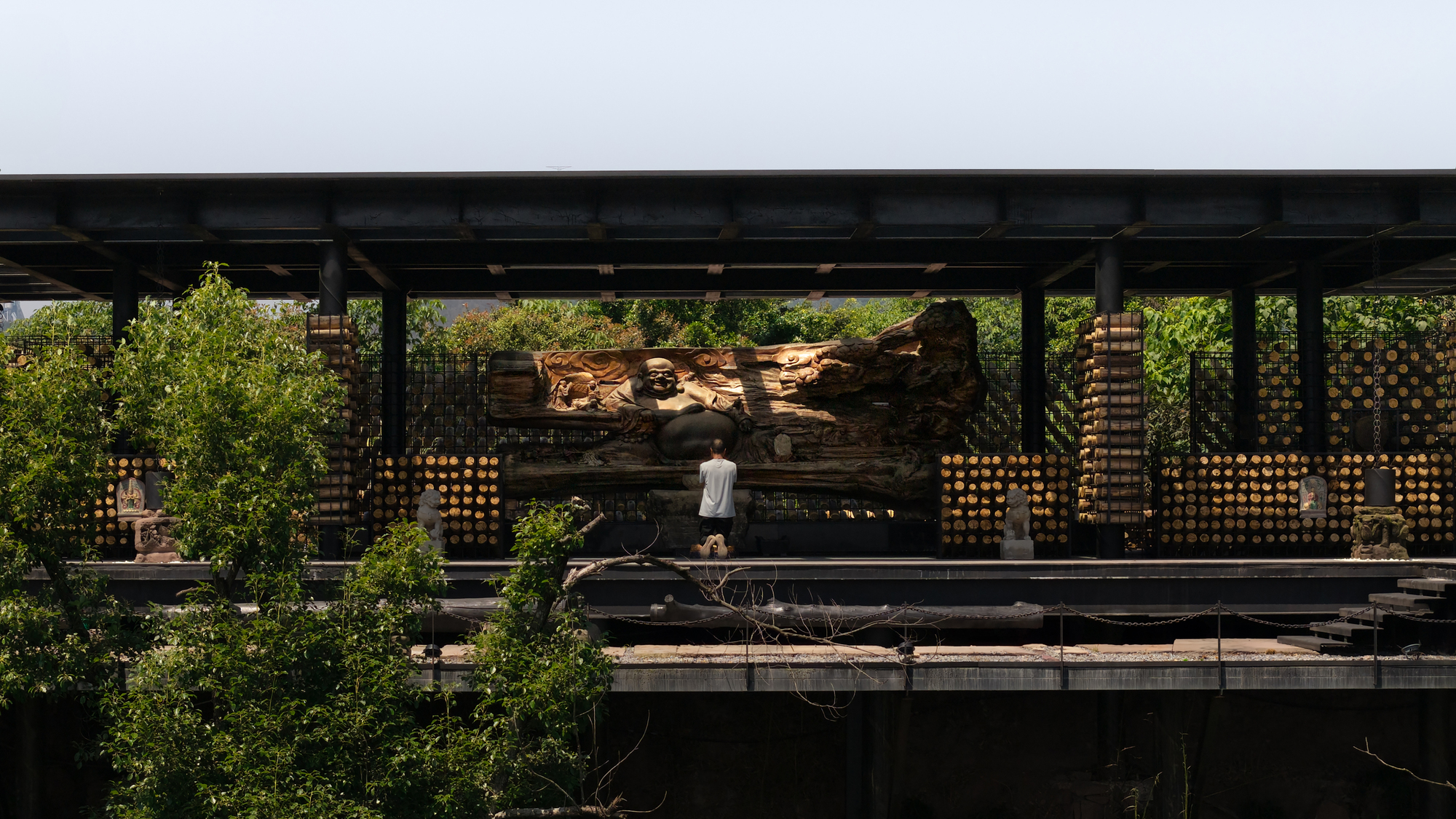 Visitor standing before a large carved wood Buddha sculpture inside an open steel-framed pavilion with log walls, in bright daylight
