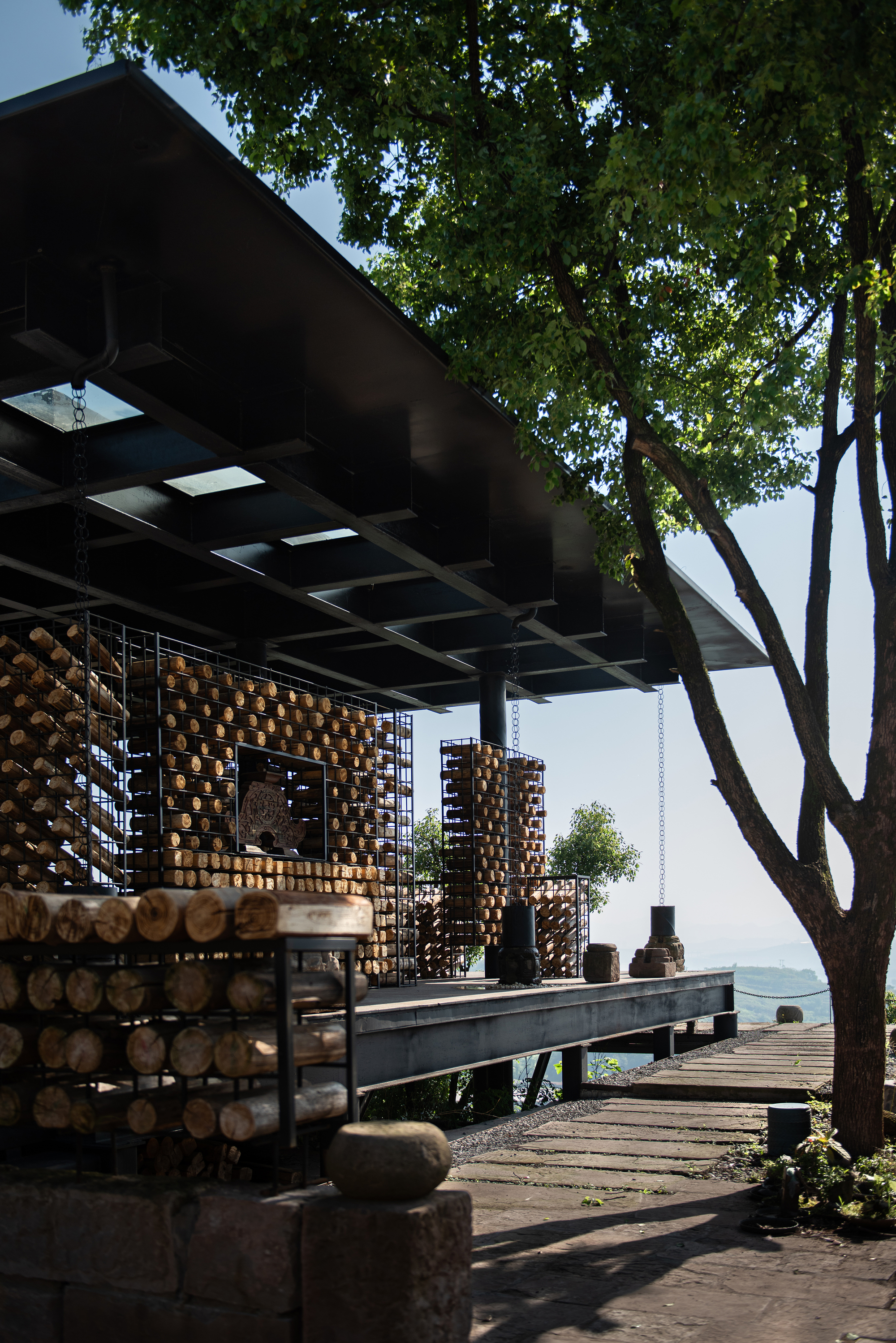Steel-framed pavilion with a dark flat roof and cages of stacked log ends, seen beside a leafy tree above a hazy valley