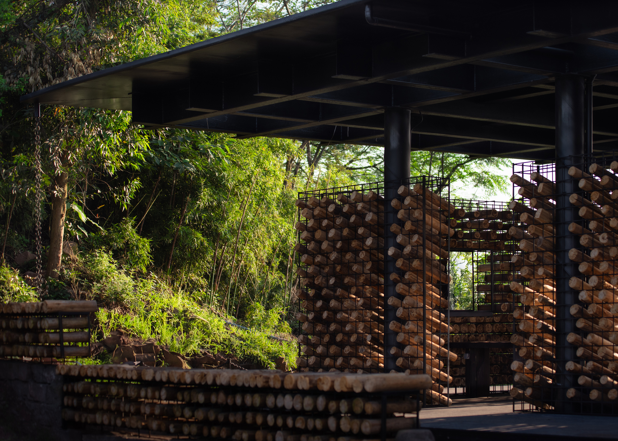 Black steel columns and wire cages filled with stacked logs under a flat roof, beside a bamboo-covered slope in warm light