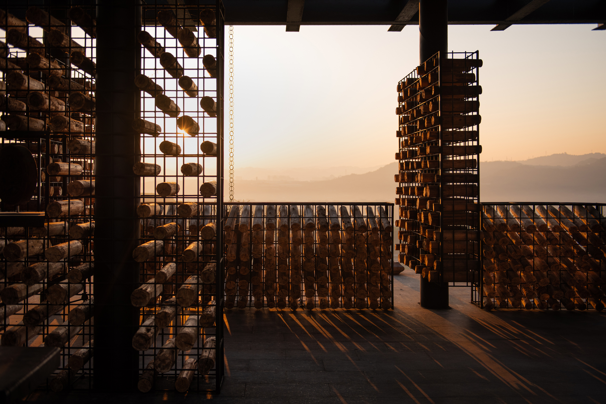Wire cages of stacked log ends silhouetted against an orange sunset over distant hills, with long shadows across the dark floor