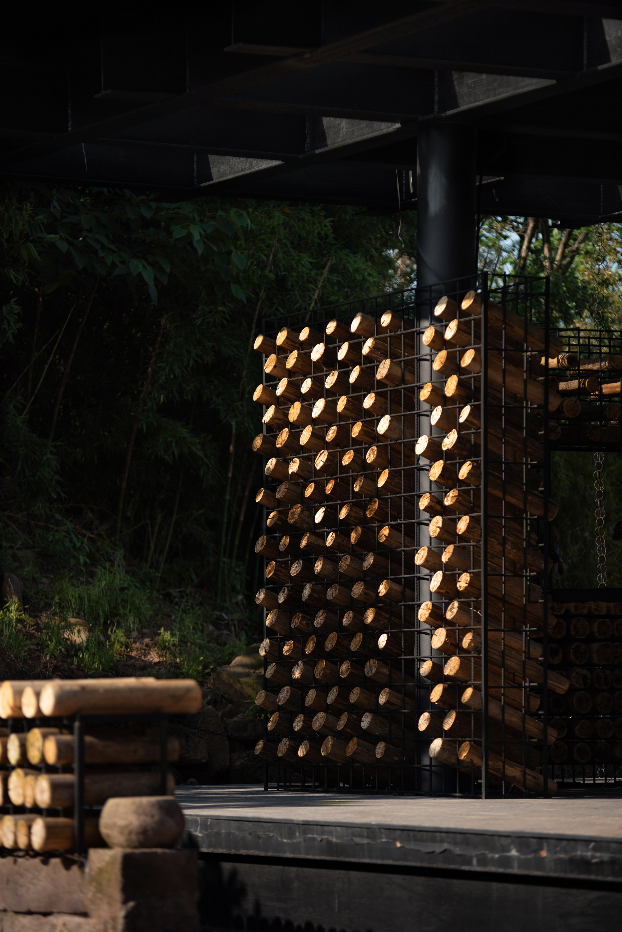 Tall wire cage of stacked logs beside a black steel column, lit by low sun against dark bamboo foliage