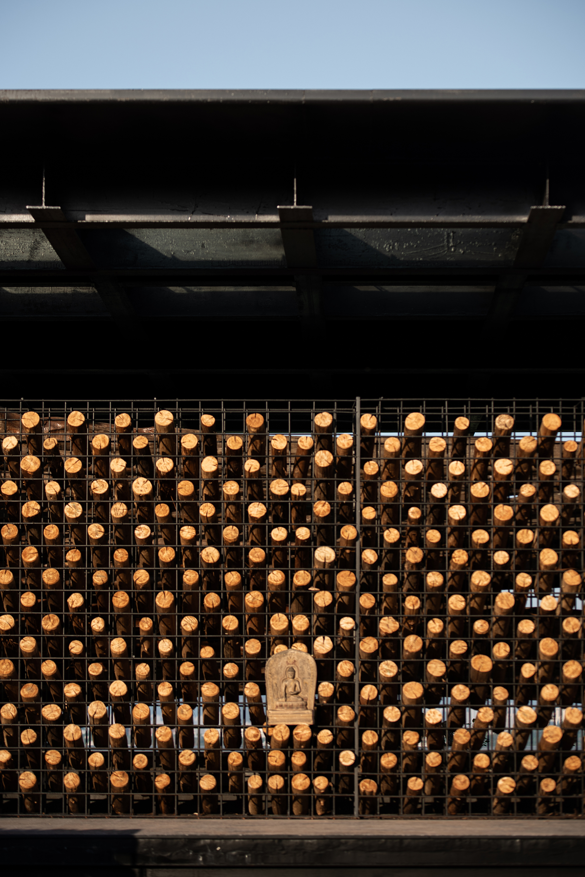 Grid of cut log ends in a steel mesh frame with a small carved Buddha relief at center under a dark roof edge