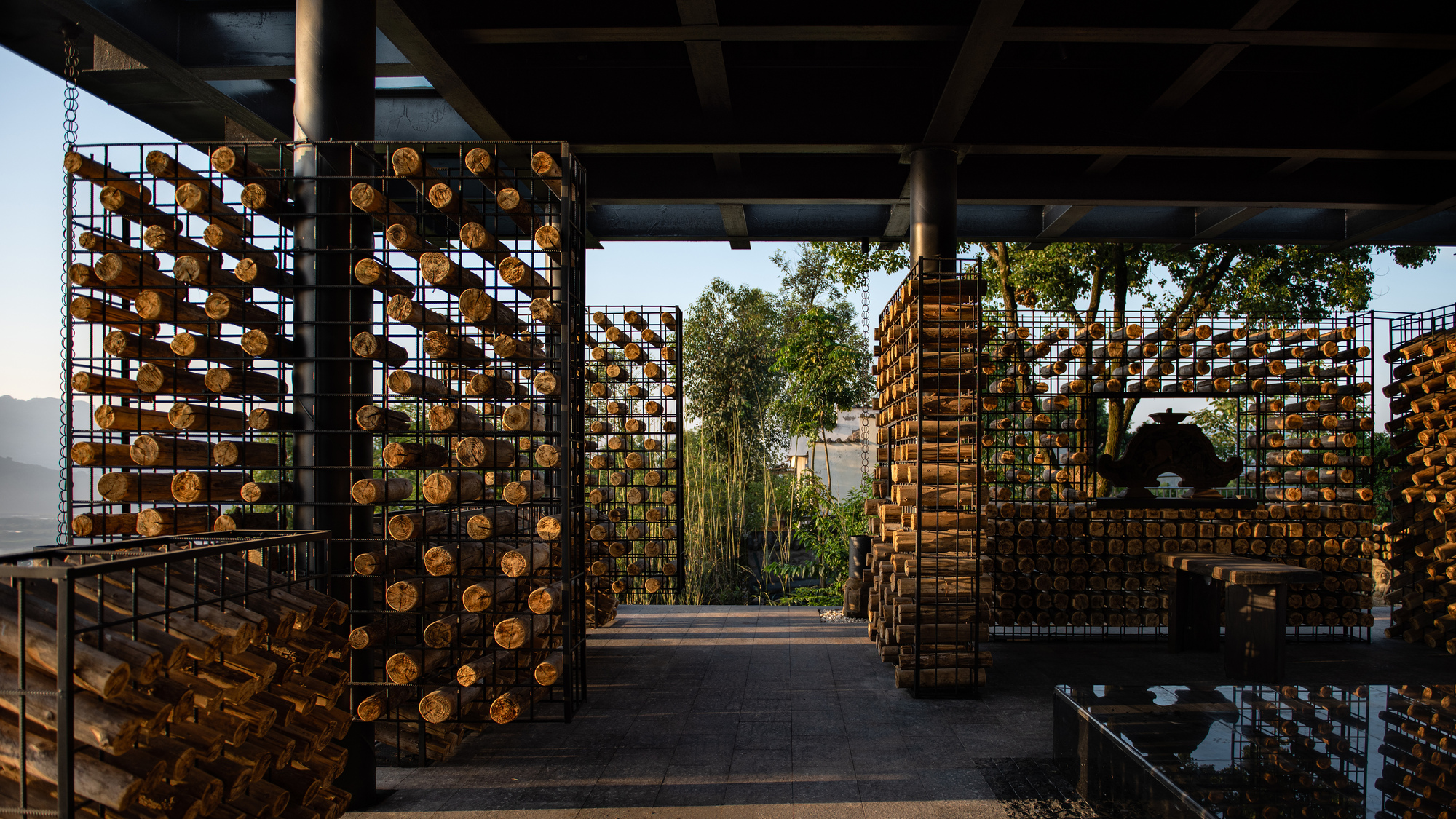 Open pavilion under a dark steel roof, with wire cages of stacked logs lit by low evening sun