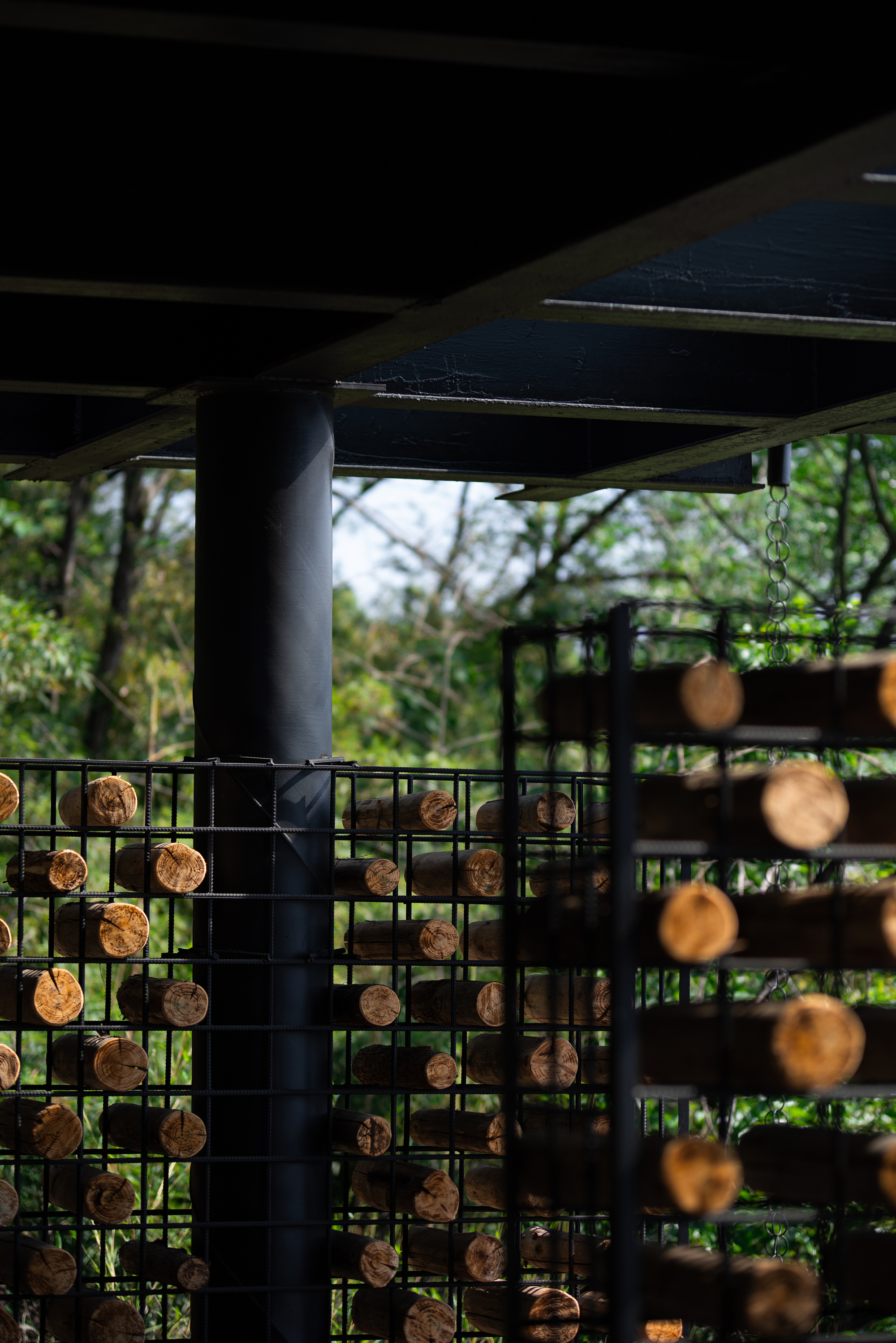 Black steel column rising beside cages of stacked log ends, with a leafy hillside glimpsed behind