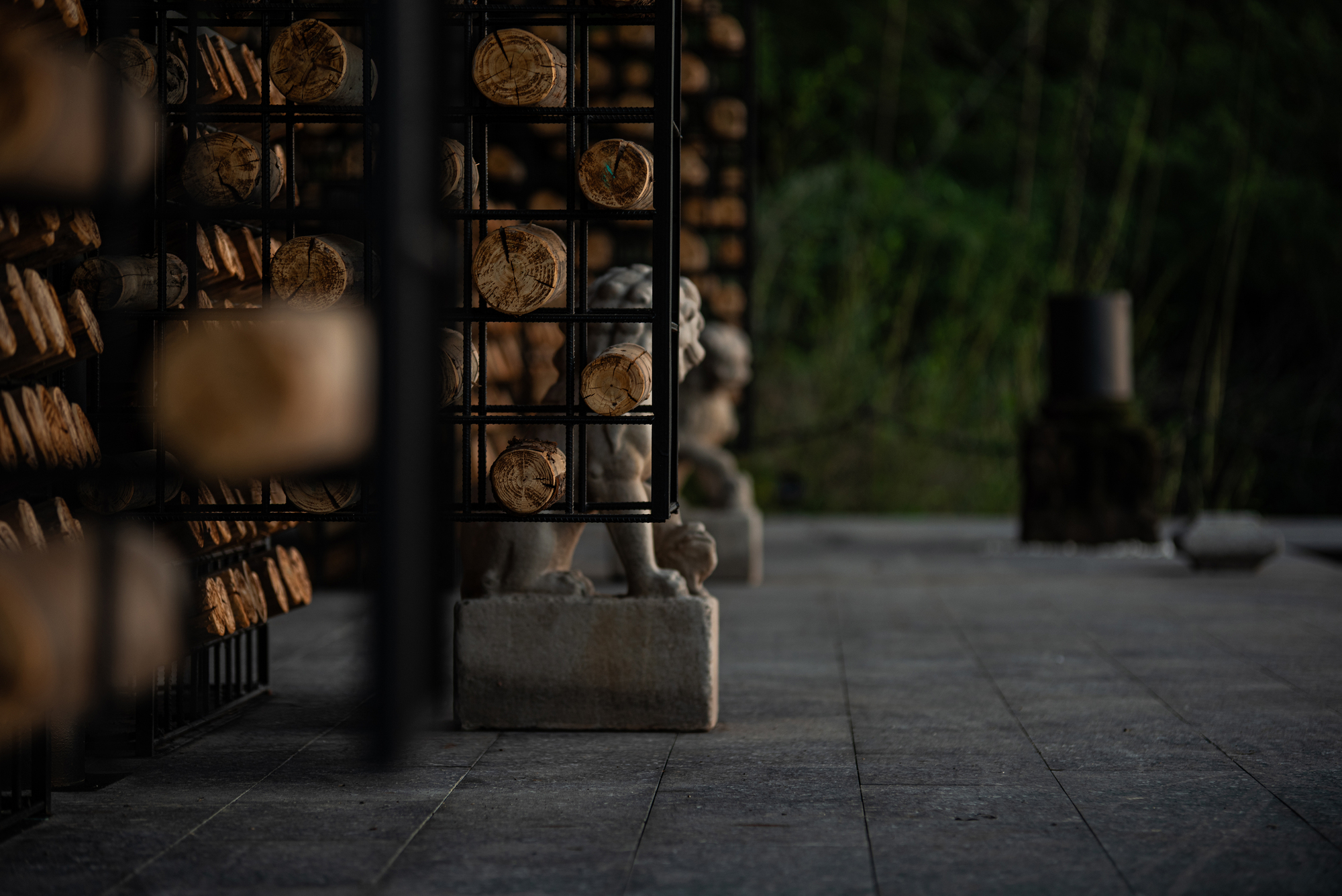 Stone lion statue on a plinth seen between log-filled steel cages, on a paved floor in dim light