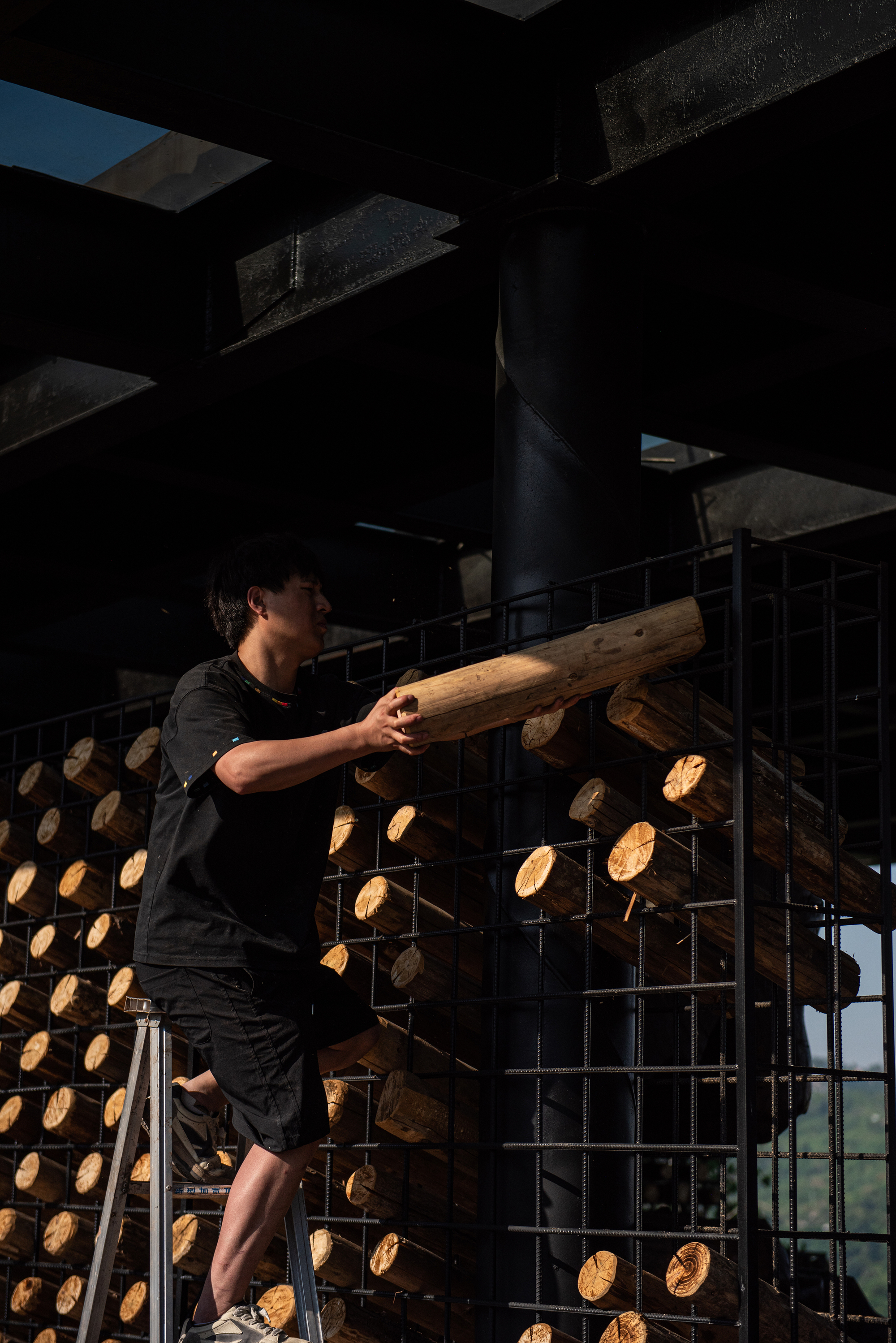Worker standing on a stepladder slides a peeled log into a steel grid under the roof beams