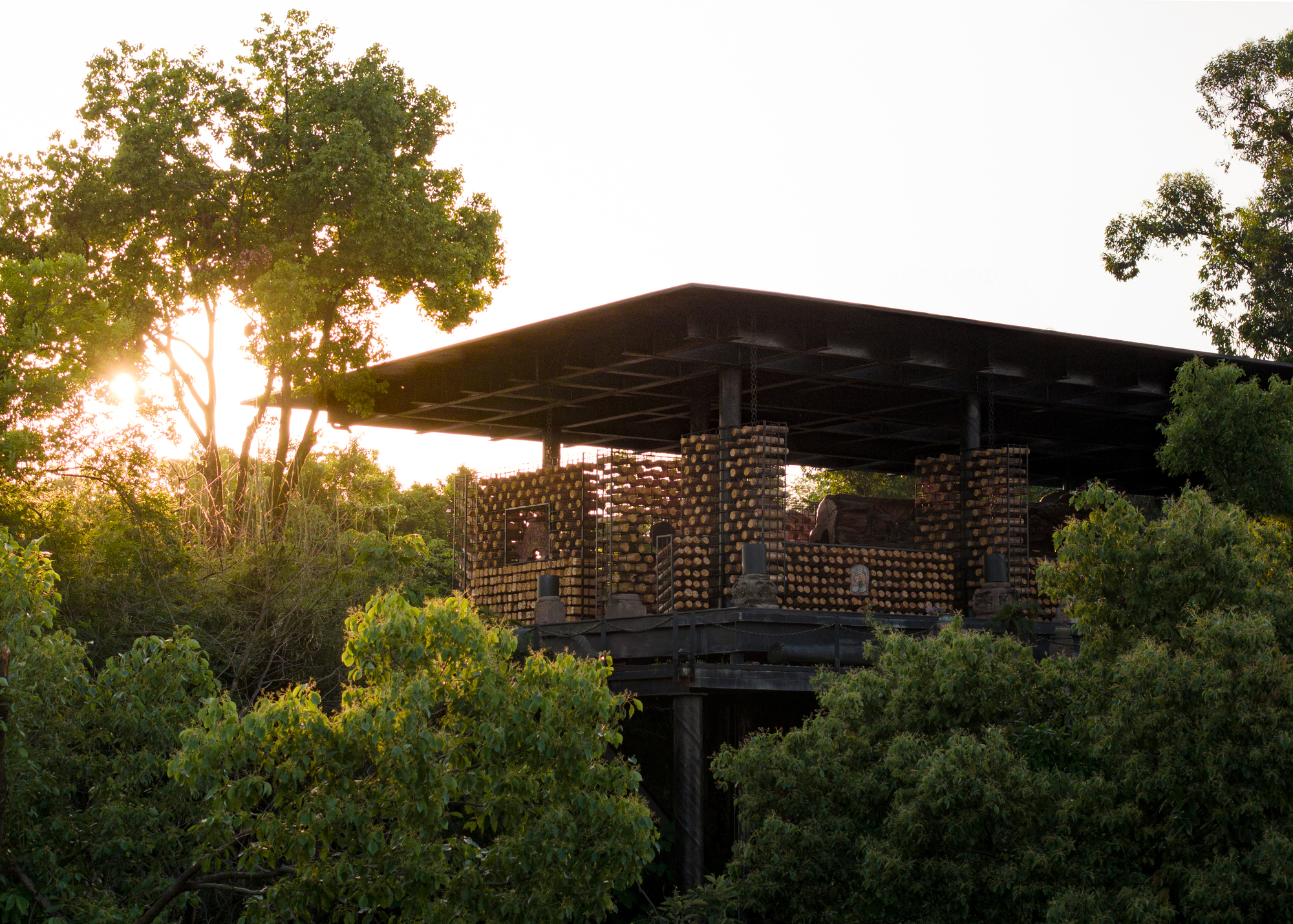 Pavilion with a flat dark roof and log-filled walls, framed by treetops and a low setting sun