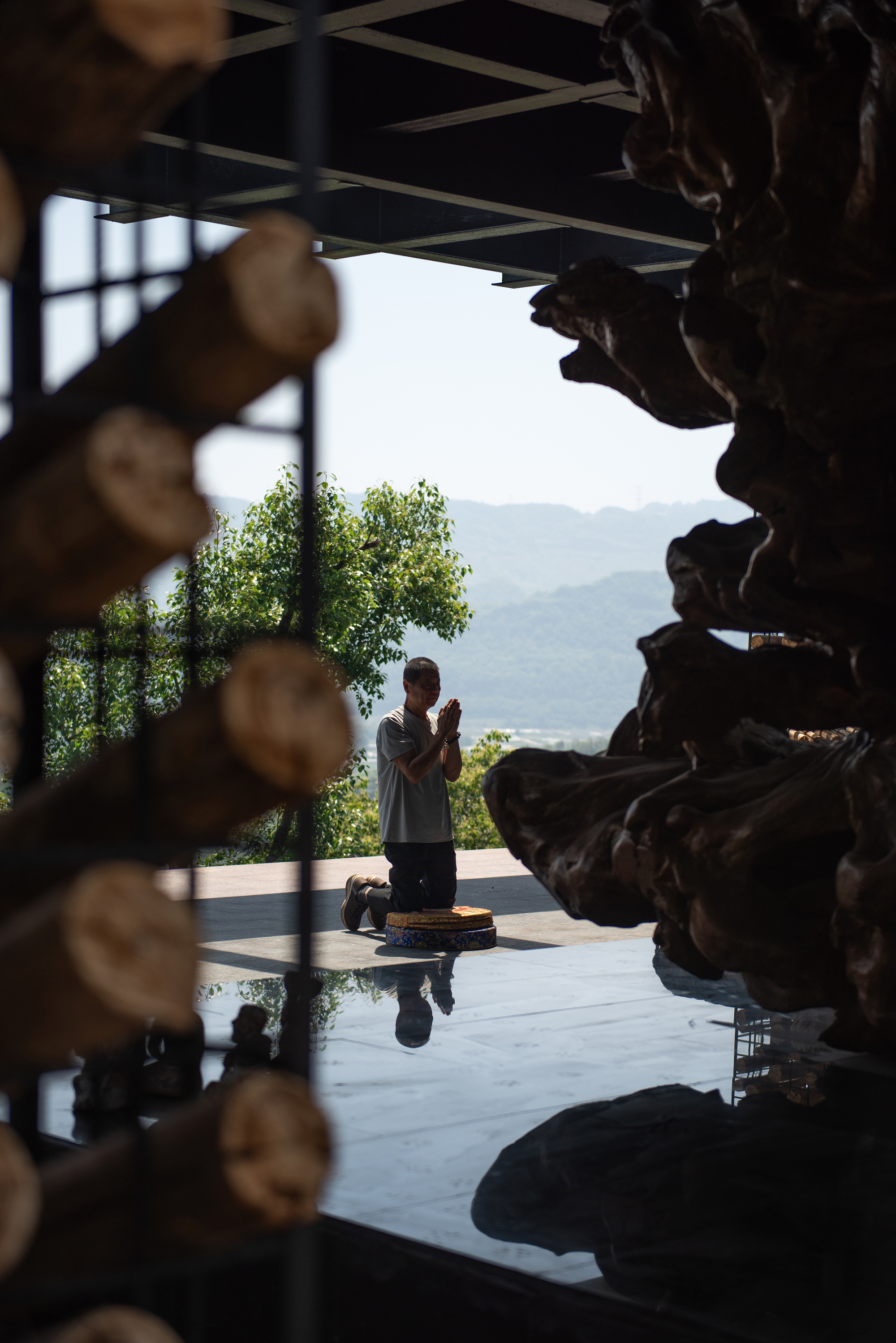 Visitor kneeling in prayer on a stone platform, framed between stacked log ends and a gnarled root sculpture, with hazy hills beyond in daylight