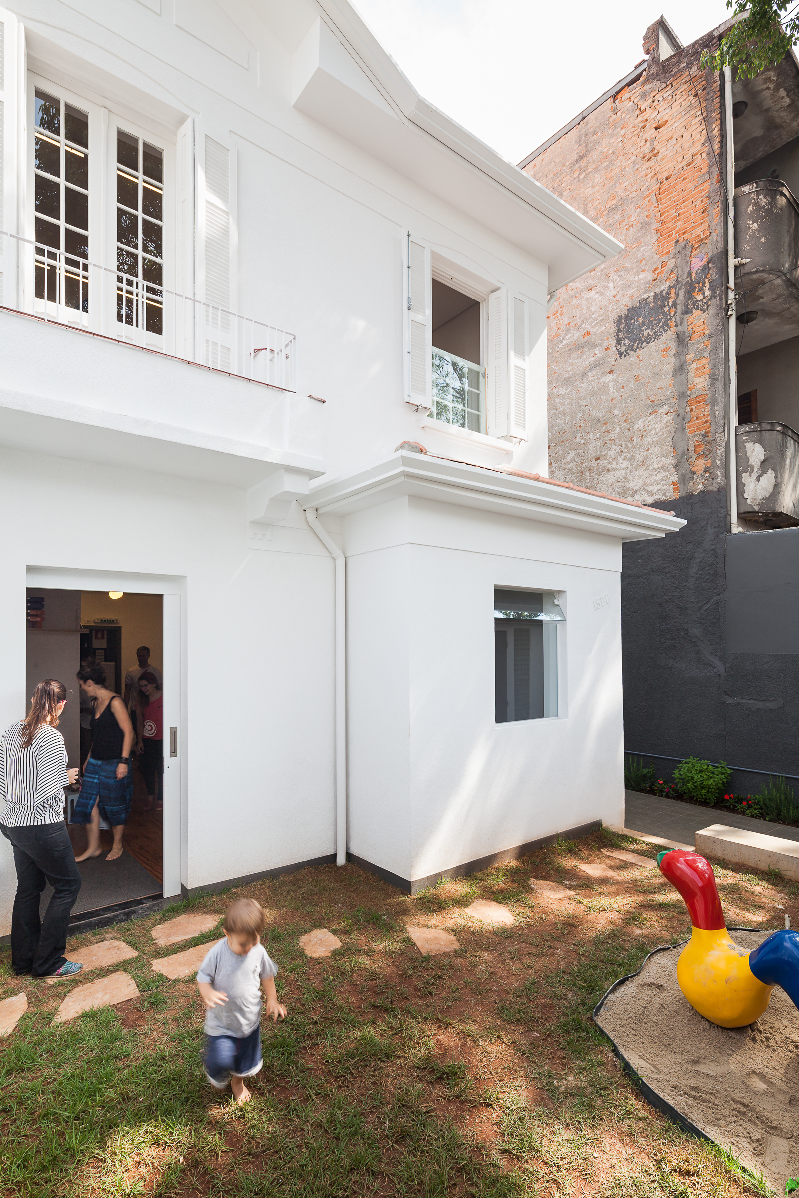 White two-storey house facade with a sliding door open to a lawn, stepping stones, a sandbox and a toddler walking in dappled sunlight
