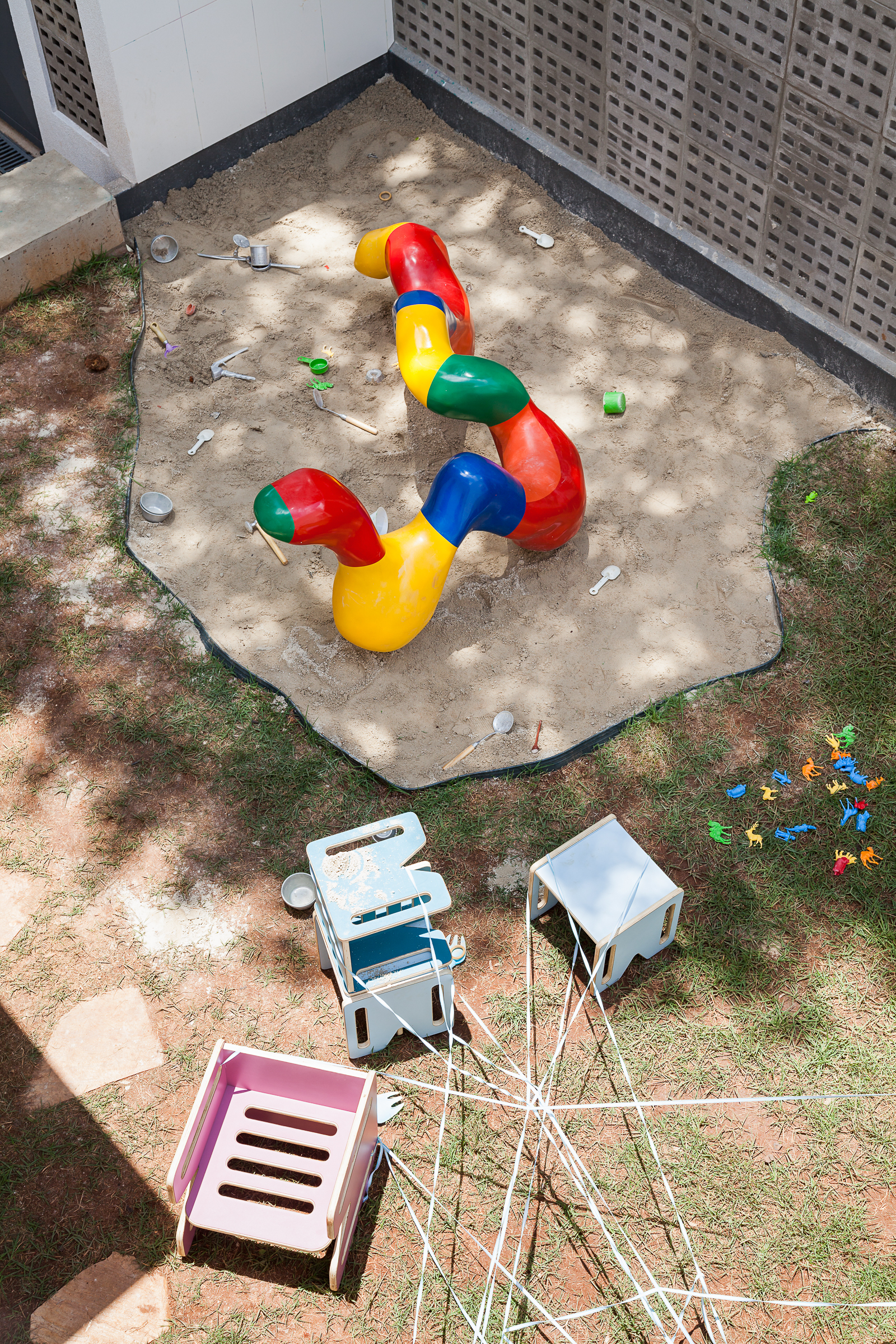 Top-down view of a sandpit with a curved red, yellow and blue play tube, scattered toys and small chairs in dappled sunlight