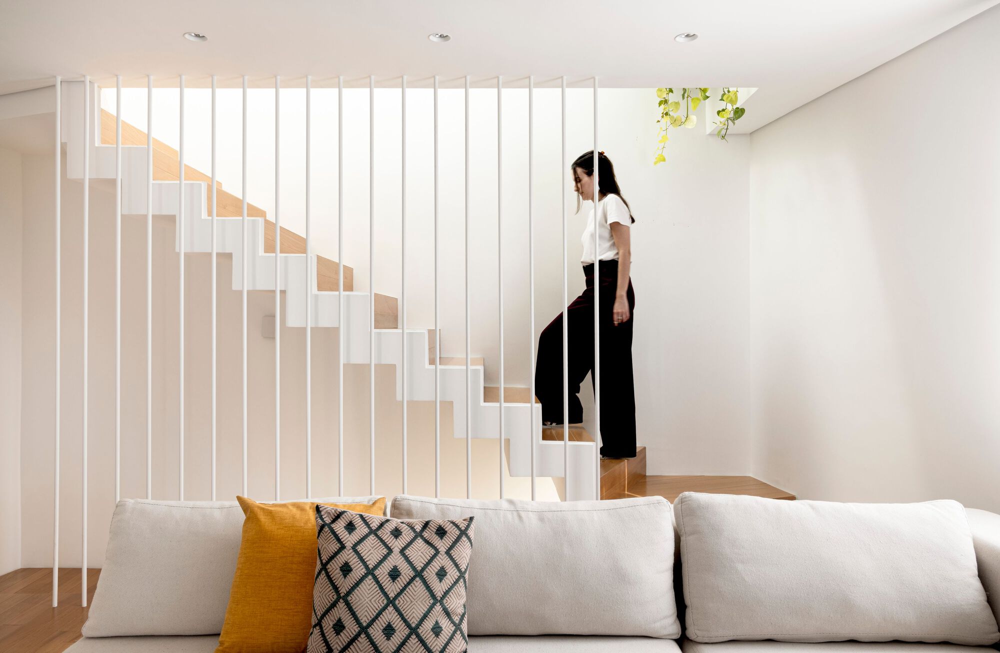 Woman climbing a white stair with thin vertical balusters above a sofa with mustard and patterned cushions in soft interior light