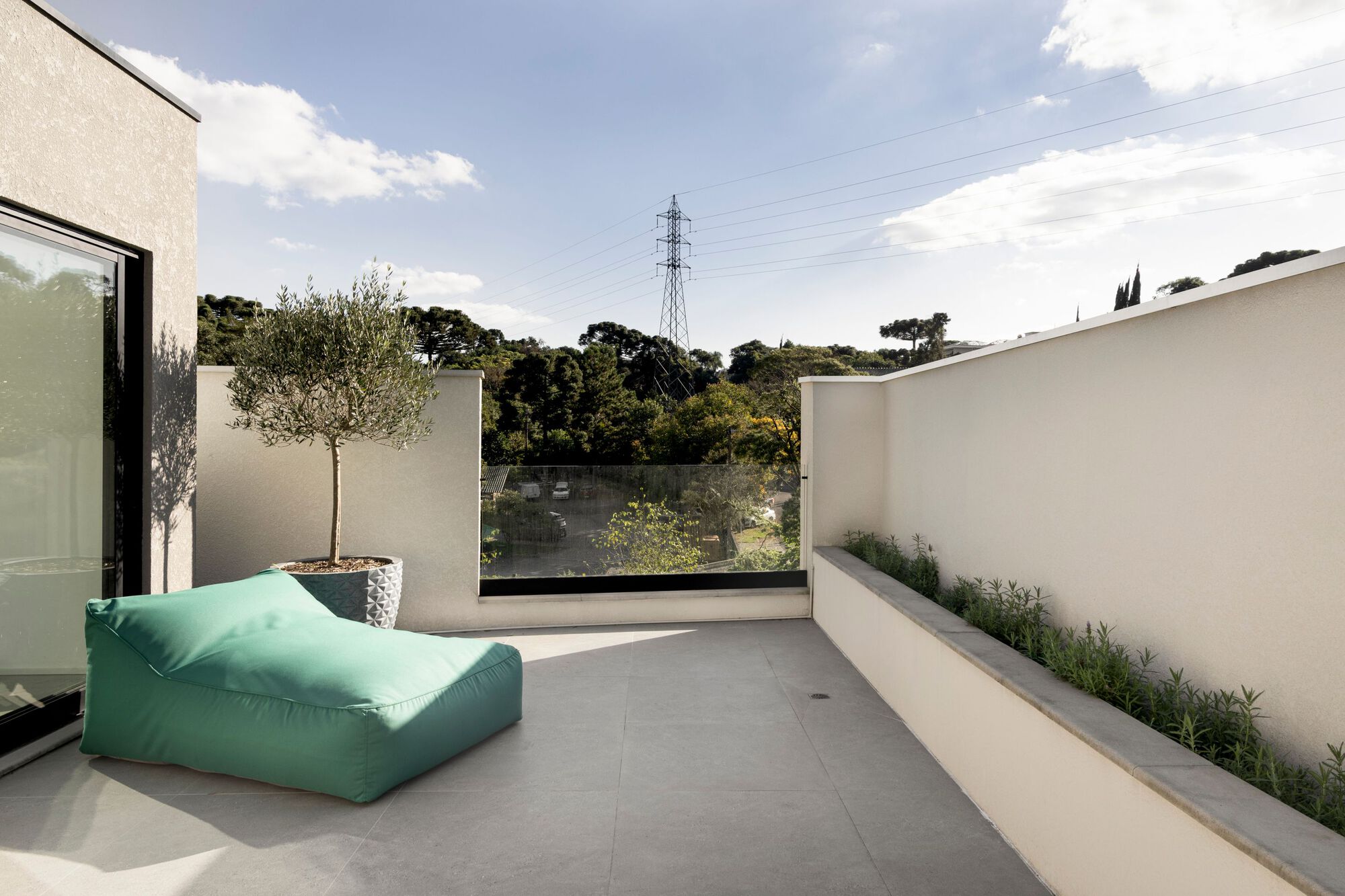 Rooftop terrace with a potted olive tree, green lounger, planter along the white wall and a glass balustrade facing distant trees