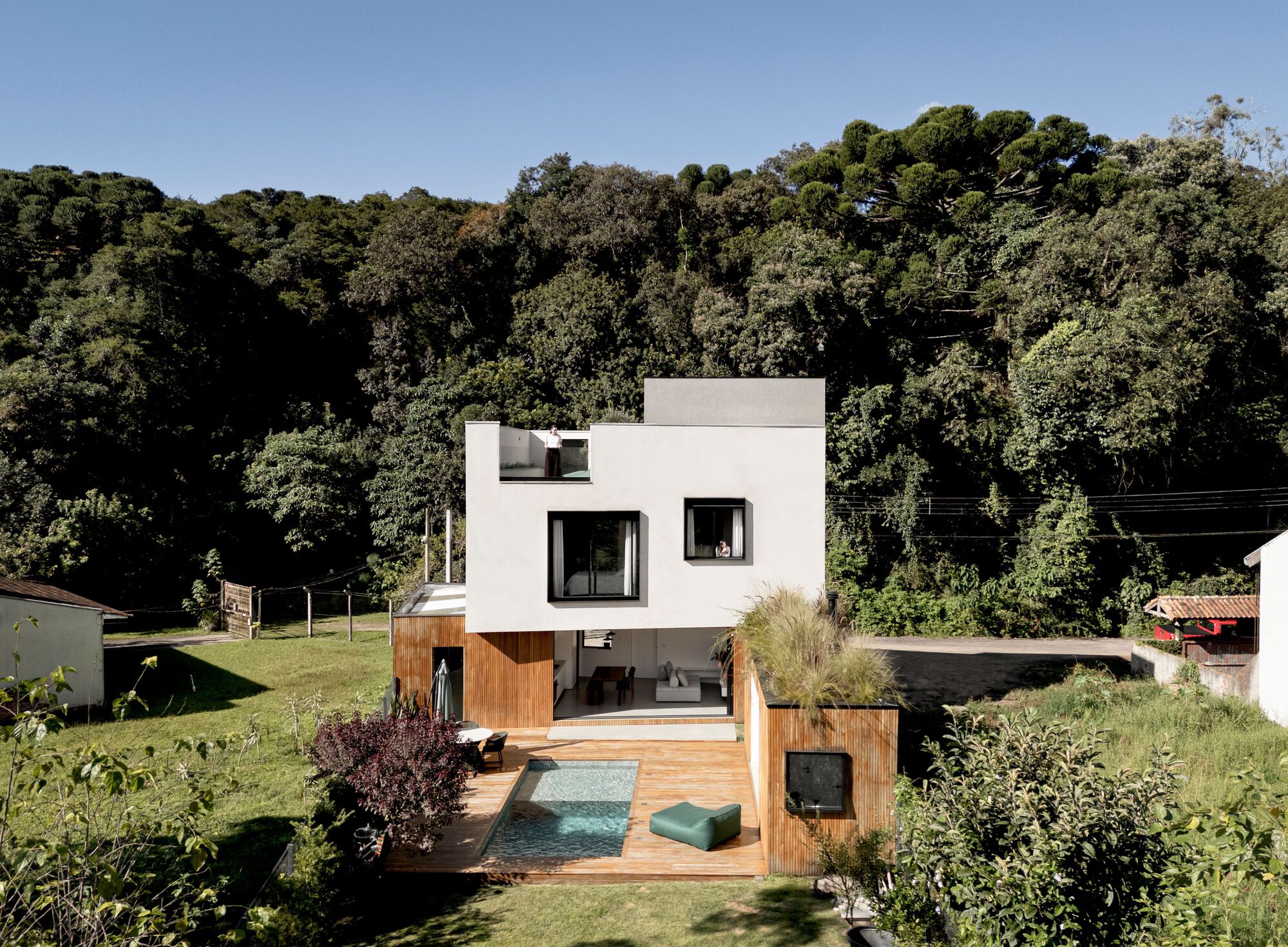 Rear view of the white house with timber base, pool and deck, set against dense forested hillside under clear blue sky