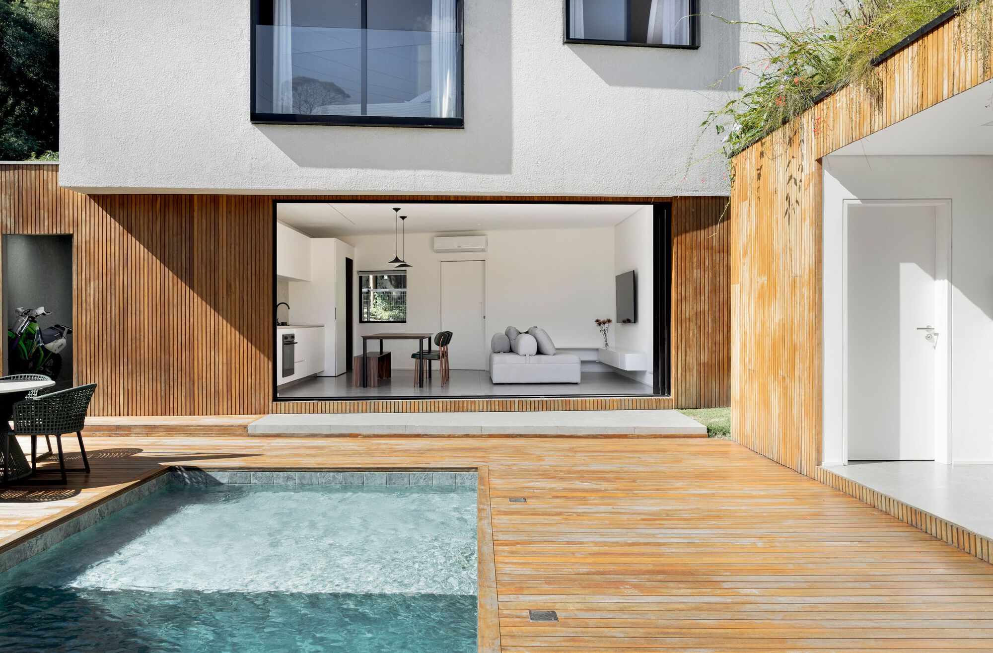 Open sliding glass wall revealing a living and kitchen space behind a slatted timber facade and pool deck in bright daylight