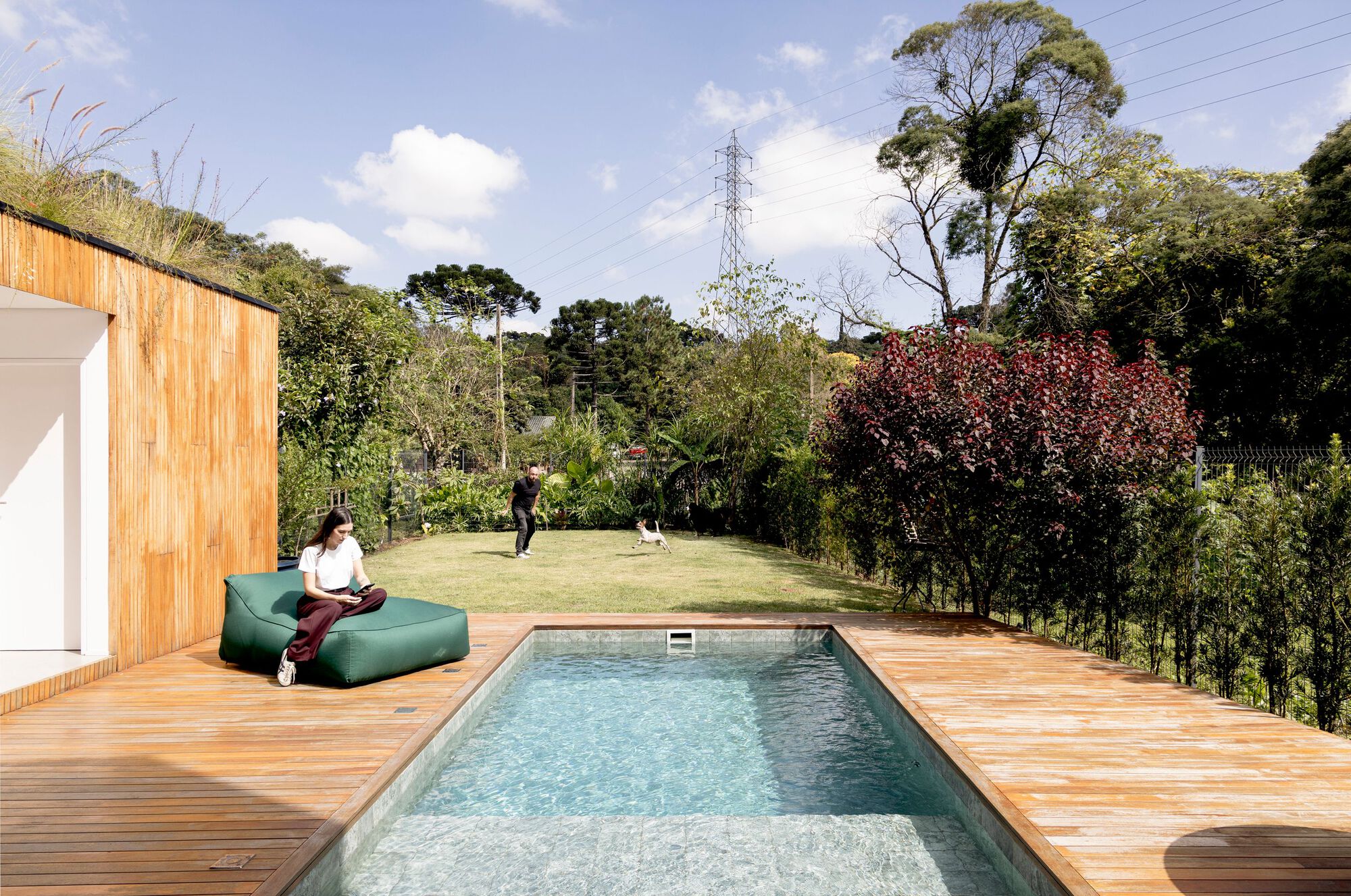 Rectangular pool set in a timber deck with a woman reading on a green pouf while a man plays with a dog on the lawn
