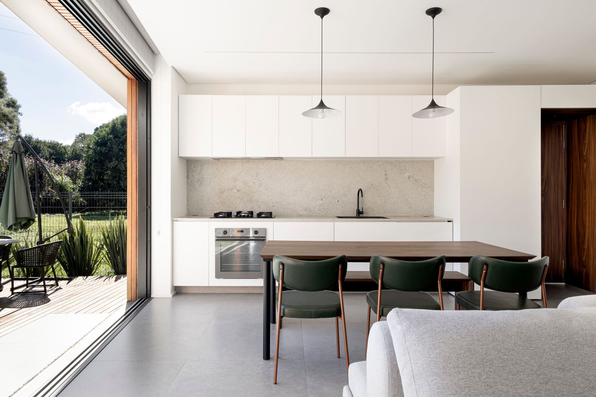 Kitchen and dining area with white cabinetry, stone backsplash, walnut table and green chairs beside an open glass wall onto the deck