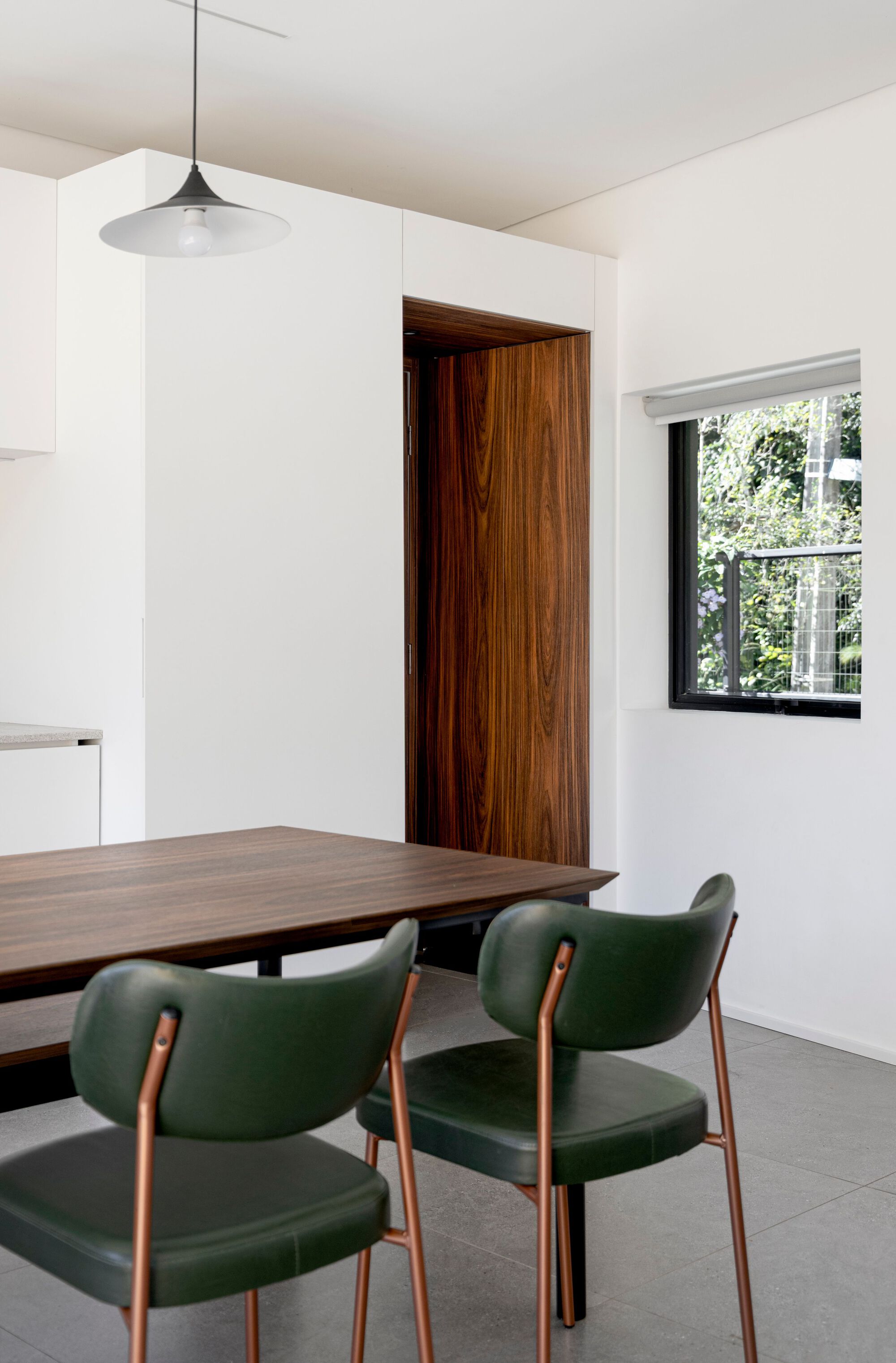 Walnut-lined door recess beside white cabinetry, with a dark timber table and green leather chairs with copper legs near a garden window