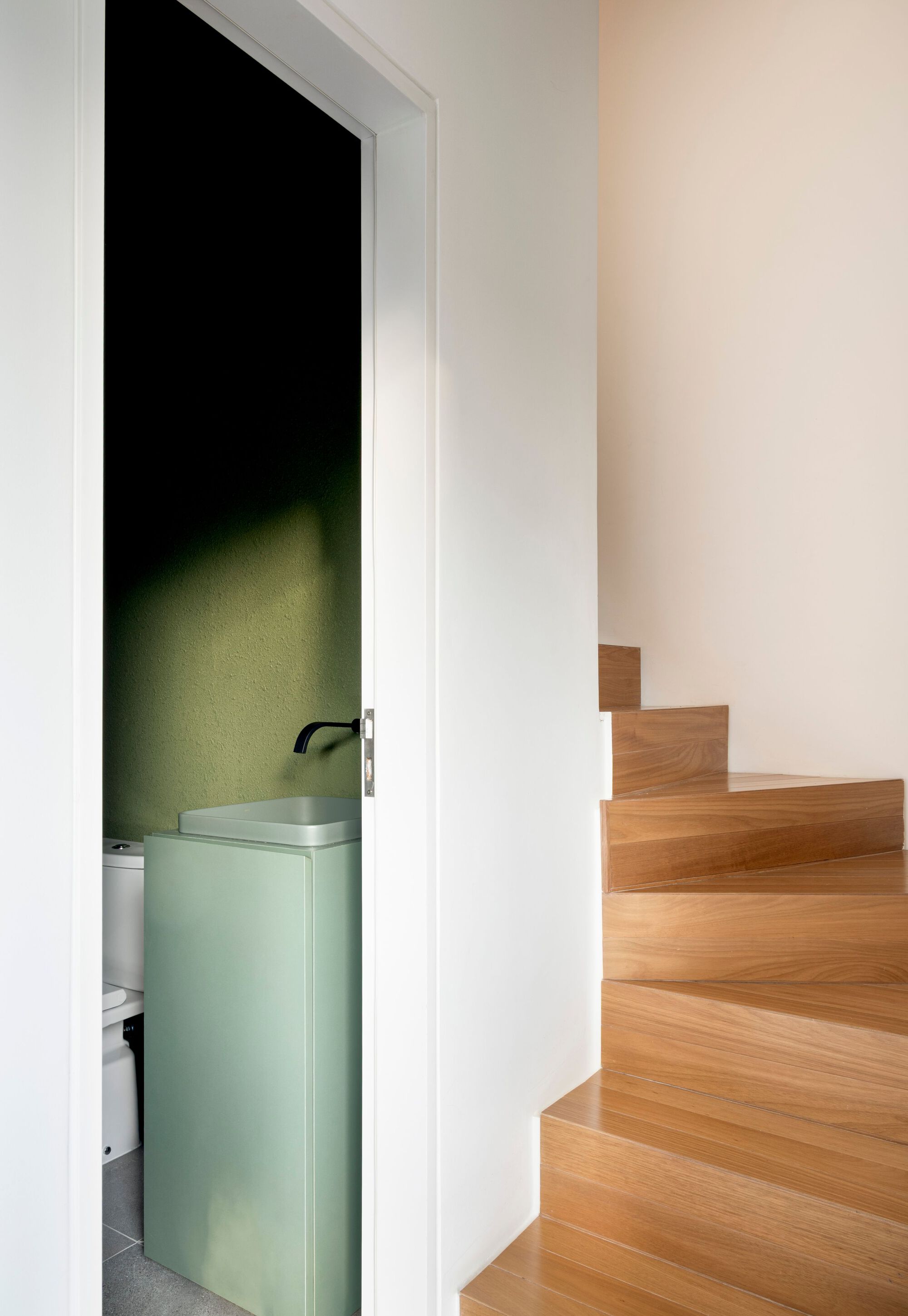 Oak stair with angled treads beside a half-open door onto a pale green vanity and moss-green wall