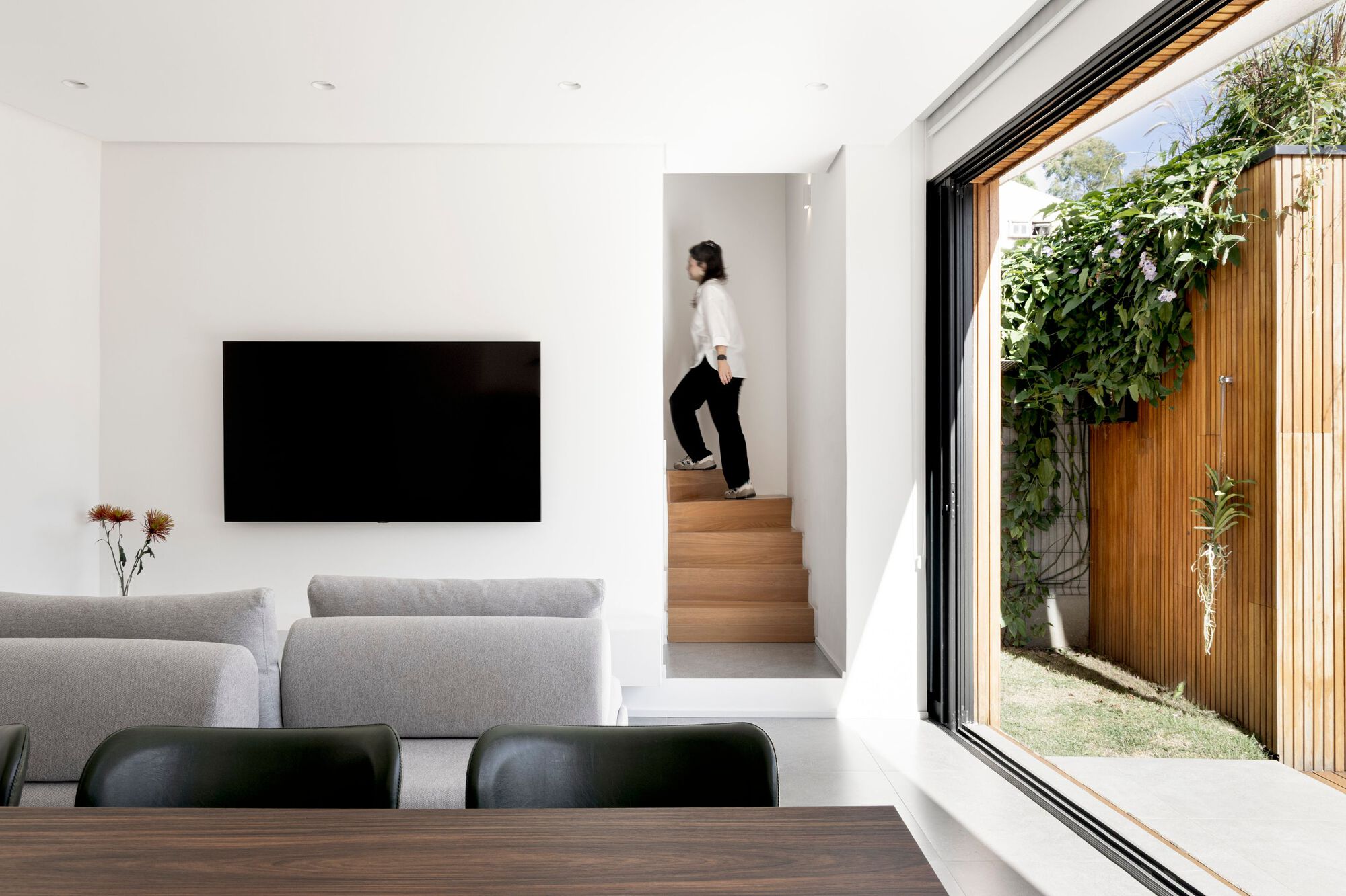 Living area opening through a sliding glass door to a timber-slatted garden wall with climbing vines, as a woman climbs the oak stair