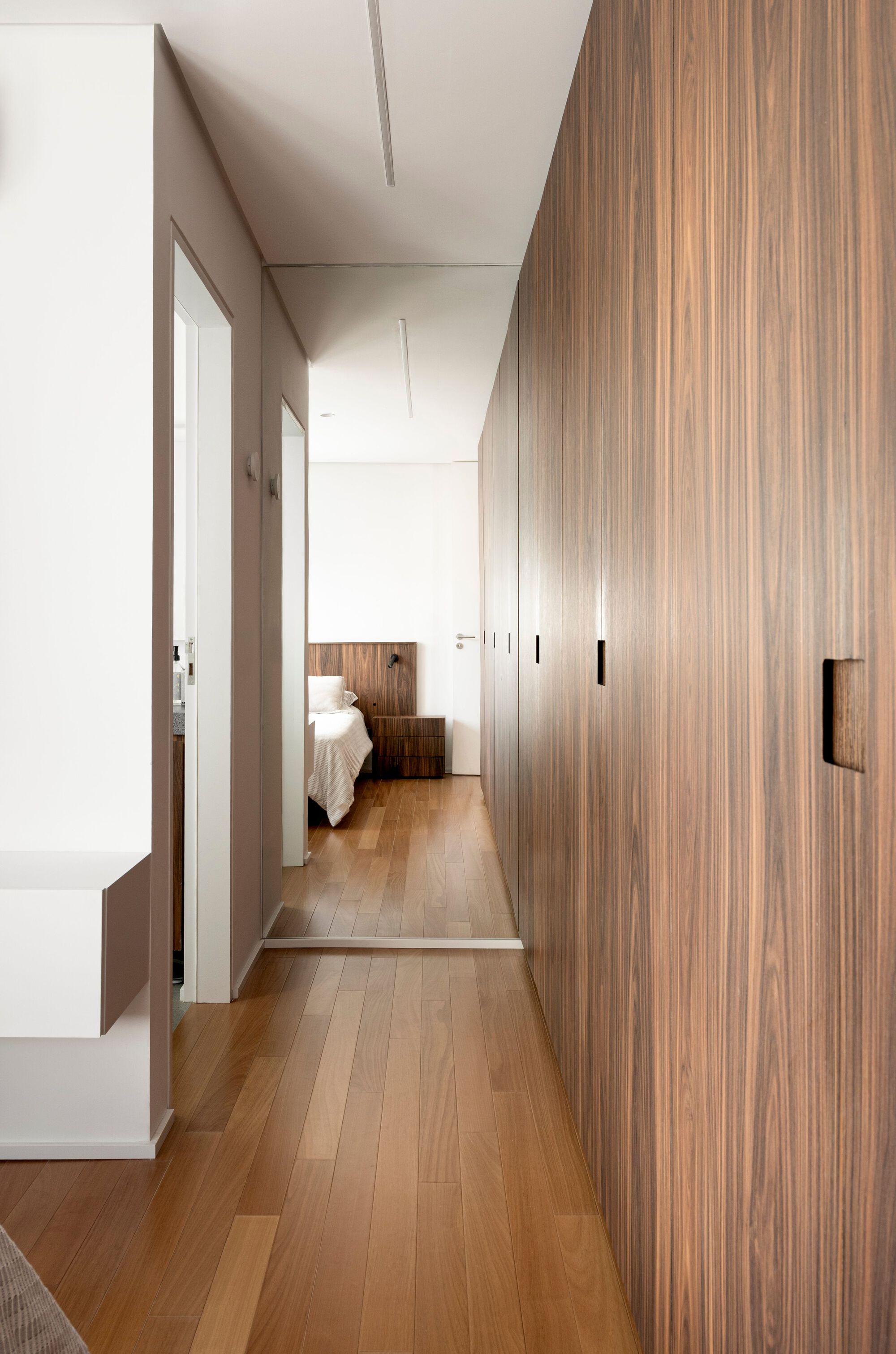 Narrow hallway lined with walnut wardrobe doors and recessed pulls, leading past oak flooring to a bedroom with a timber headboard