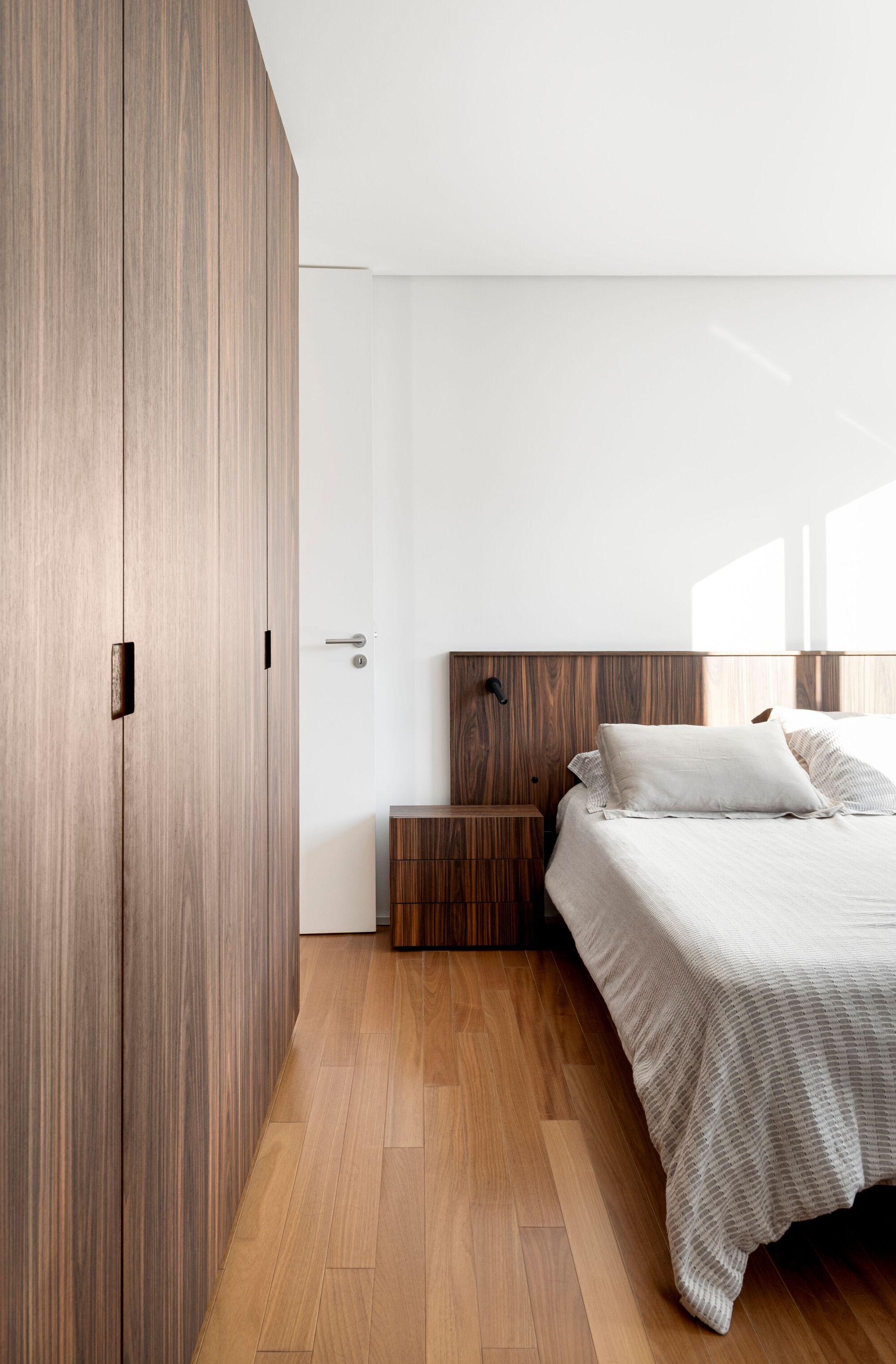 Bedroom with a walnut headboard, matching bedside drawers and tall wardrobe doors, with afternoon sun patches on the white wall